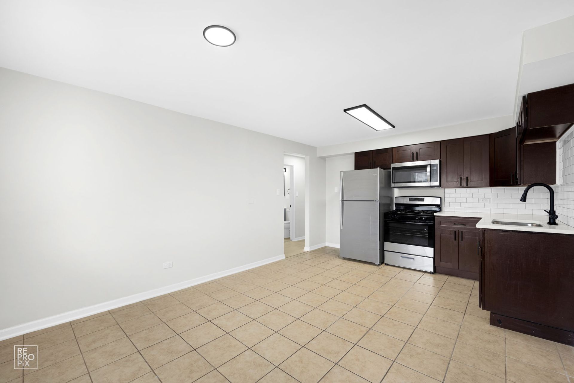Empty kitchen with dark brown cabinets, stainless steel appliances, and beige tile floor.