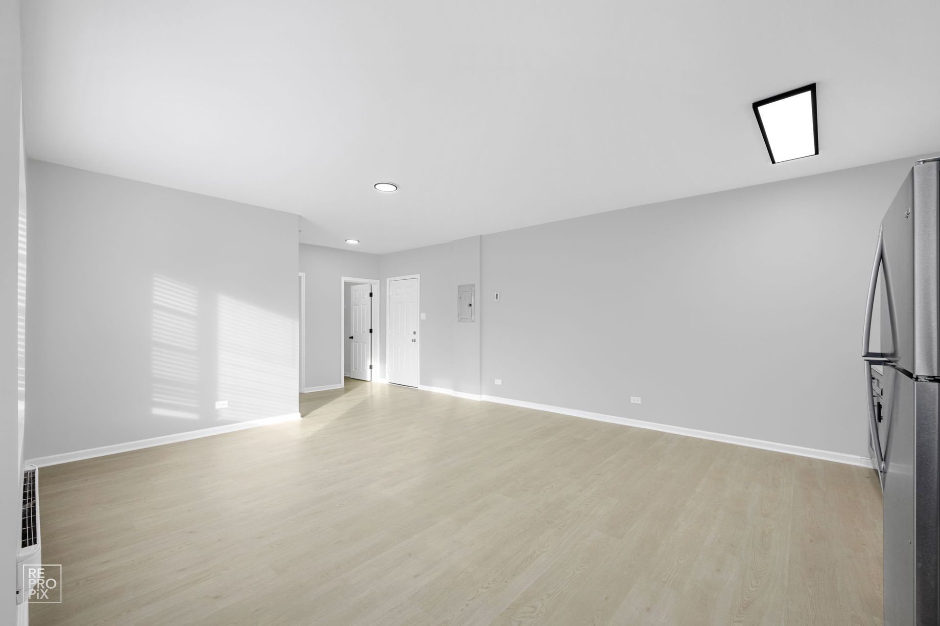 Empty living room with light gray walls, wood-look floors, and a stainless steel refrigerator.