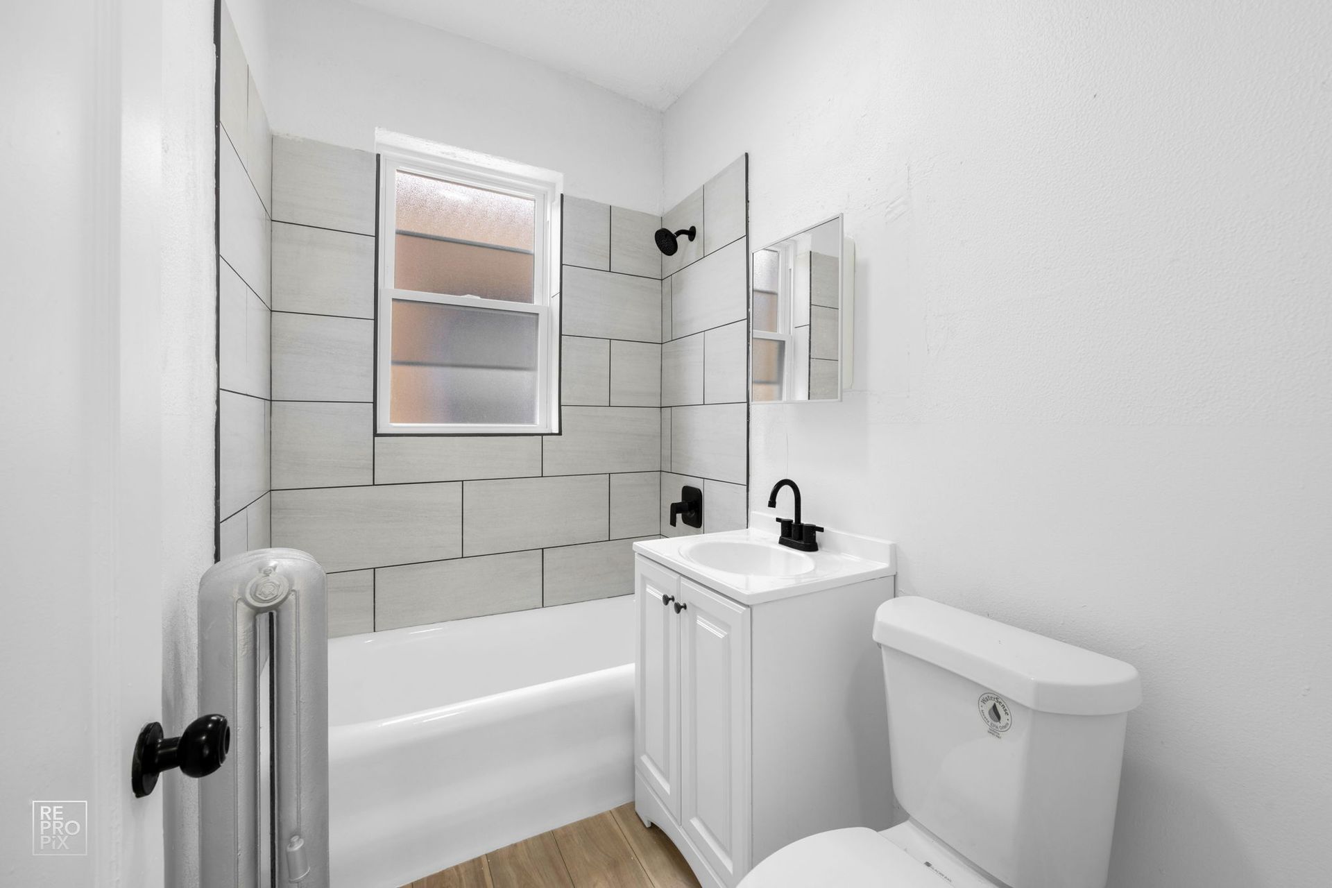 White bathroom with a bathtub, toilet, vanity, and small window. Gray tiled walls and black fixtures.