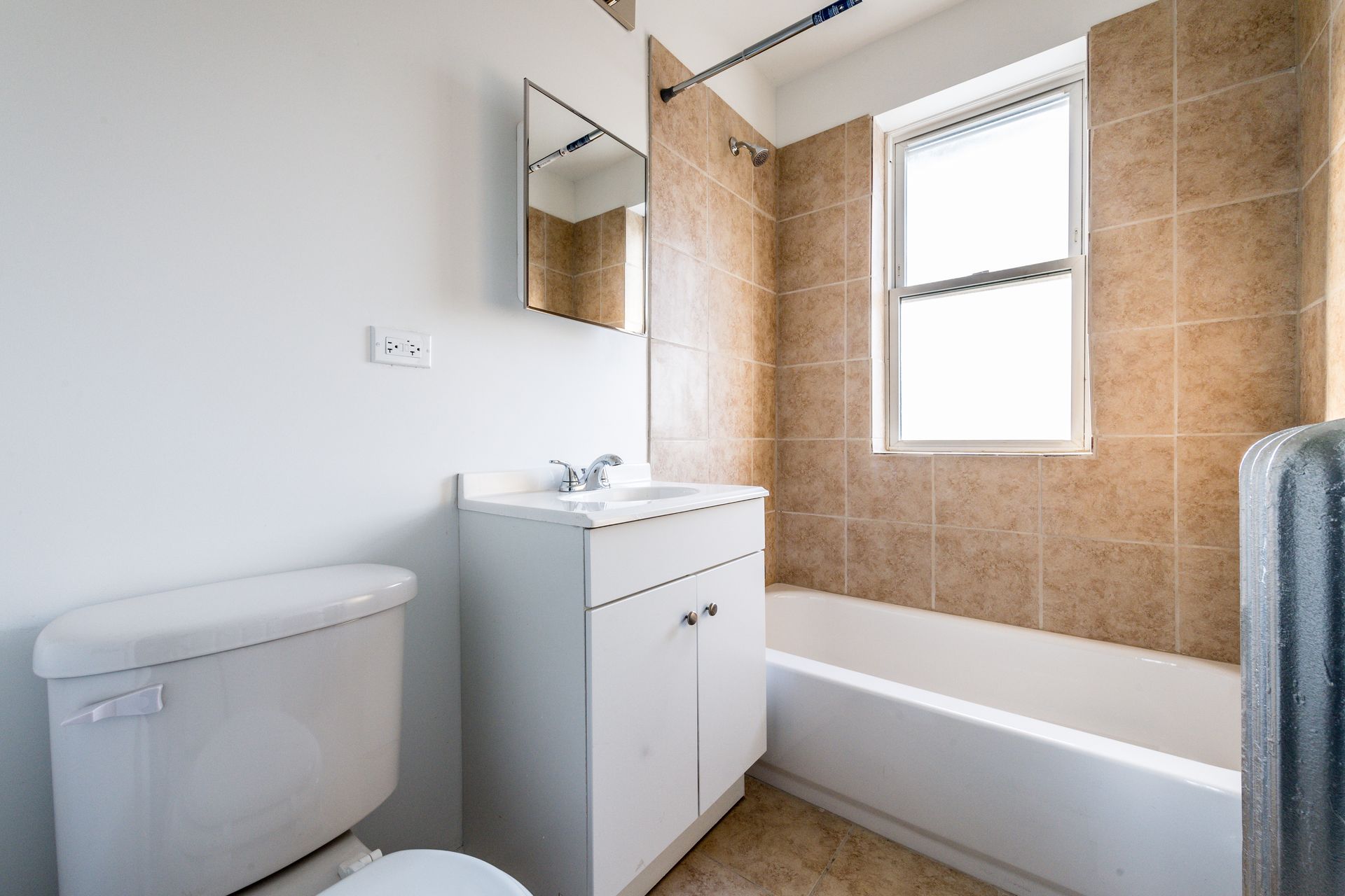Bathroom with white toilet, vanity, and bathtub; tan tile wall; window with natural light.