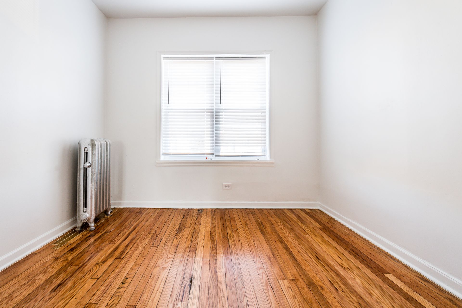 Empty room with wood floor, white walls, radiator, and window with blinds.
