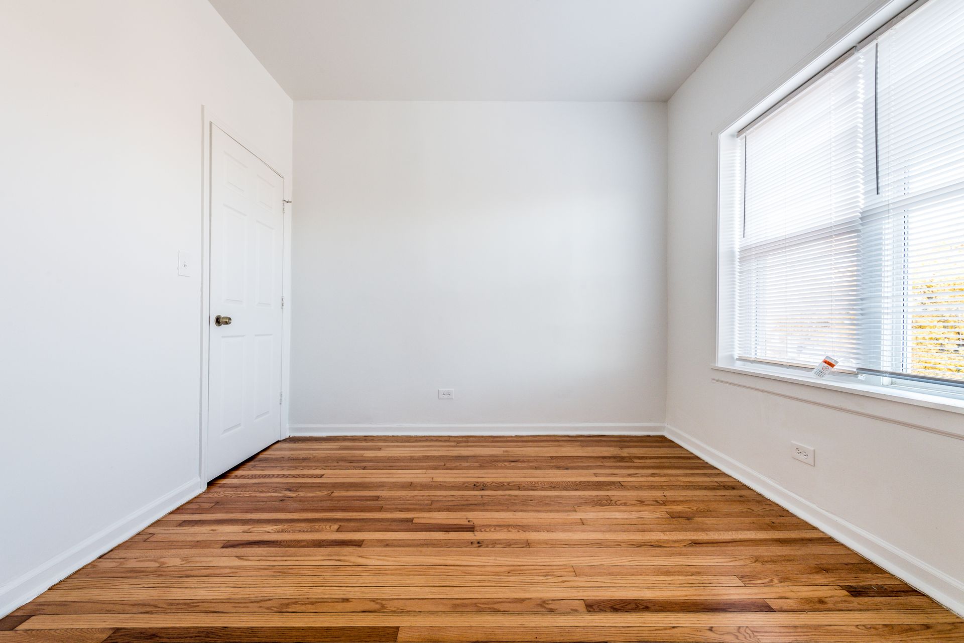 Empty room with hardwood floors, white walls, a door, and a window with blinds.