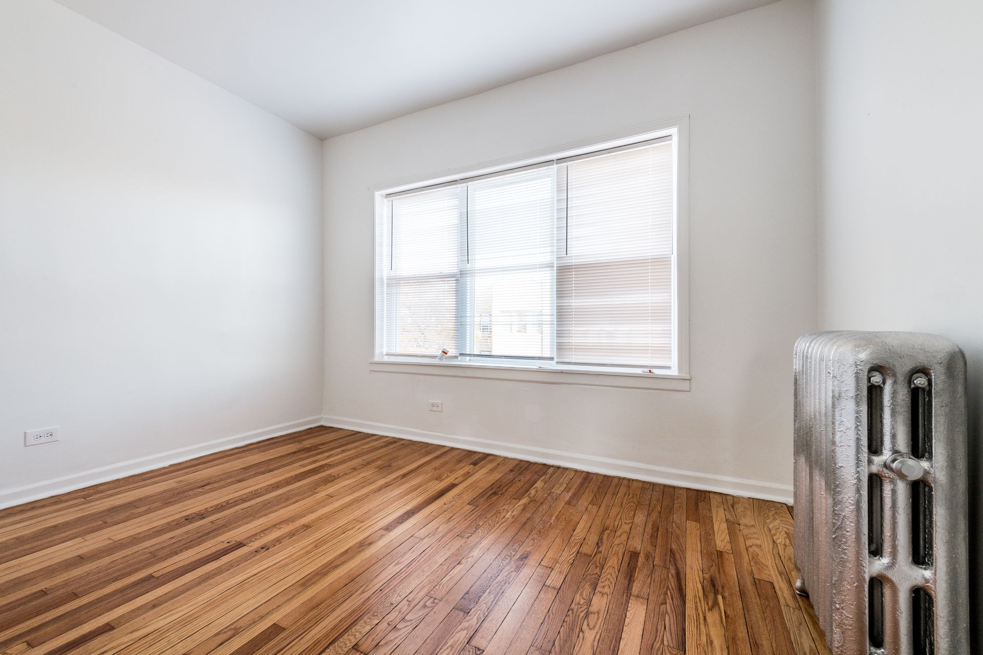 Empty room with hardwood floors, window with blinds, and a radiator.