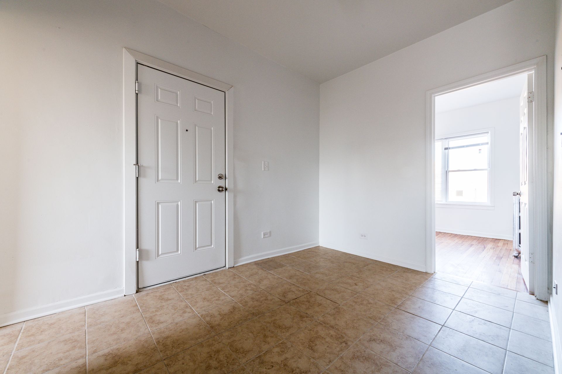 Empty room with a closed white door and a doorway to another room. Beige flooring and white walls.
