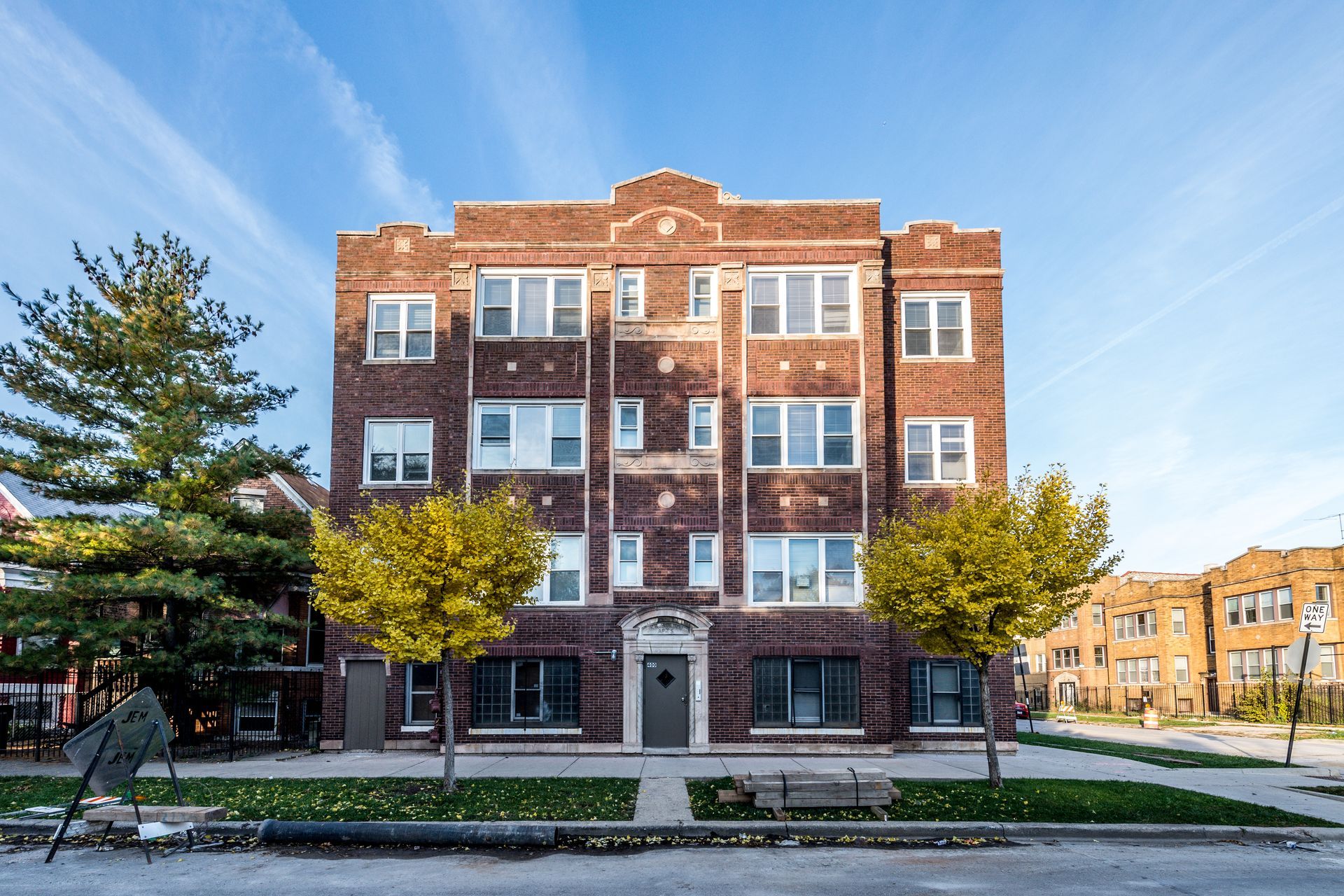 Brick apartment building with multiple stories, windows, and decorative top. Trees and street visible.
