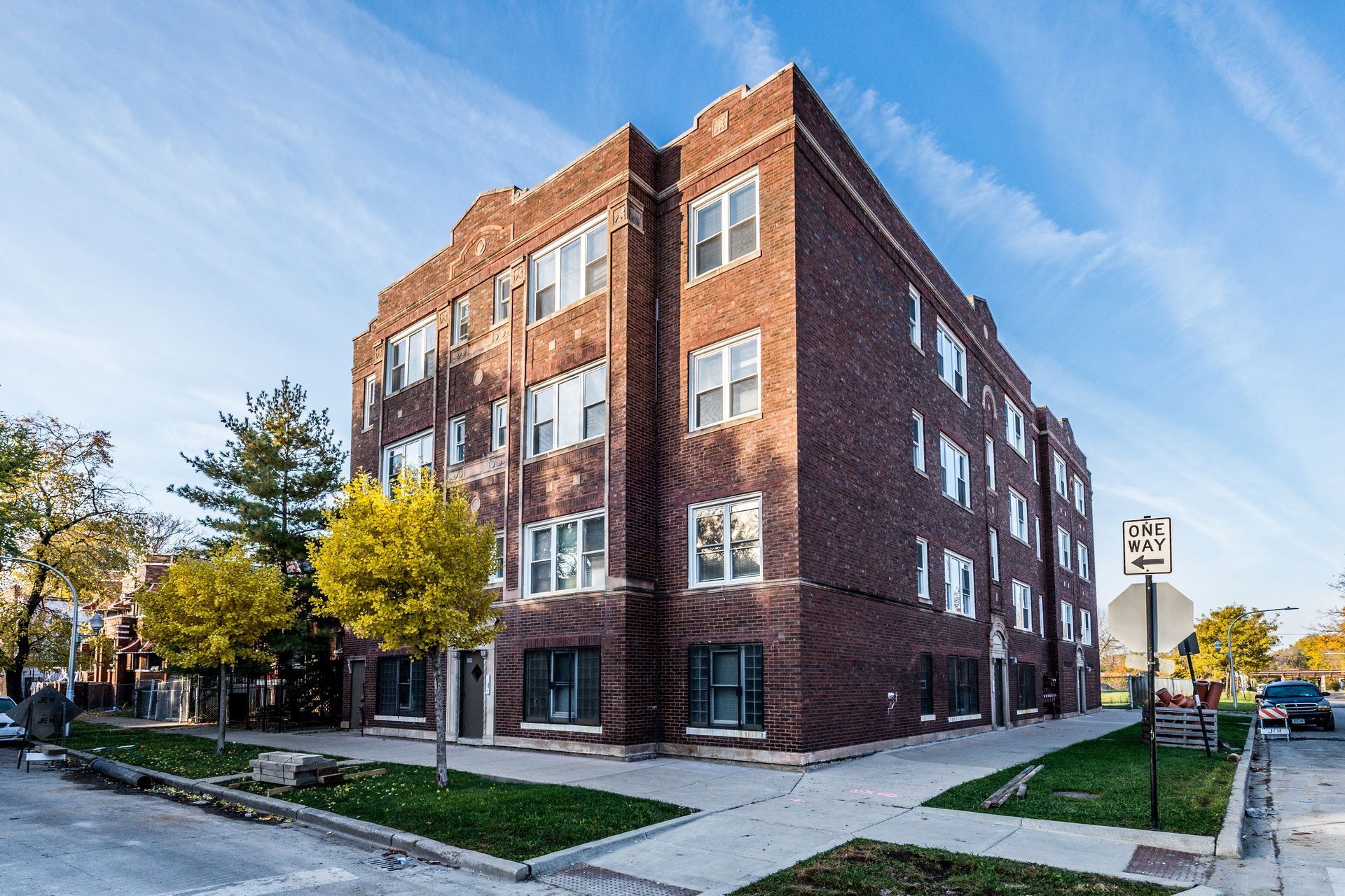 Brick apartment building on a corner lot; blue sky, trees, sidewalk, street.