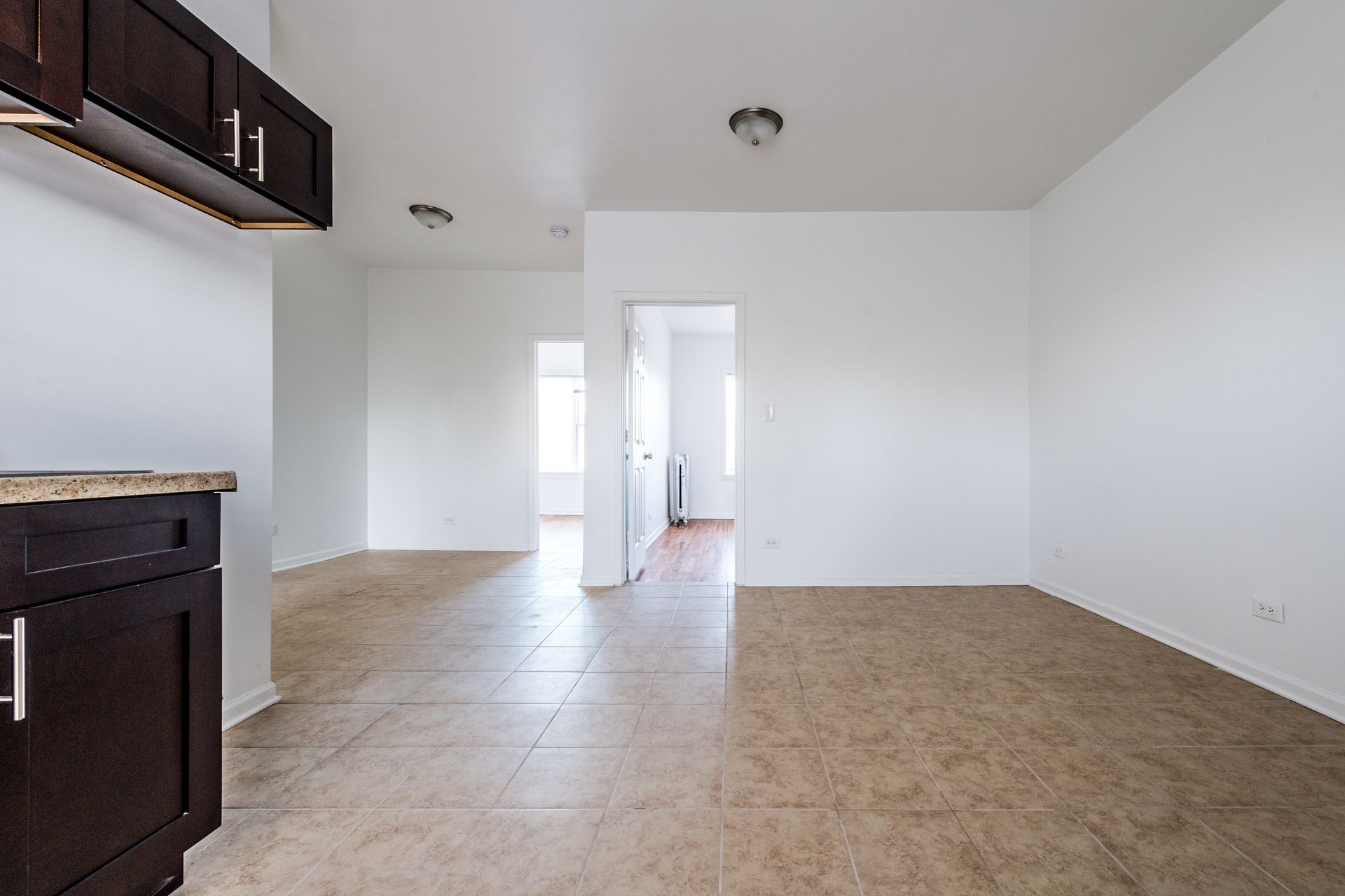 Empty kitchen and adjacent rooms with brown cabinets, tan tile floor, and white walls.