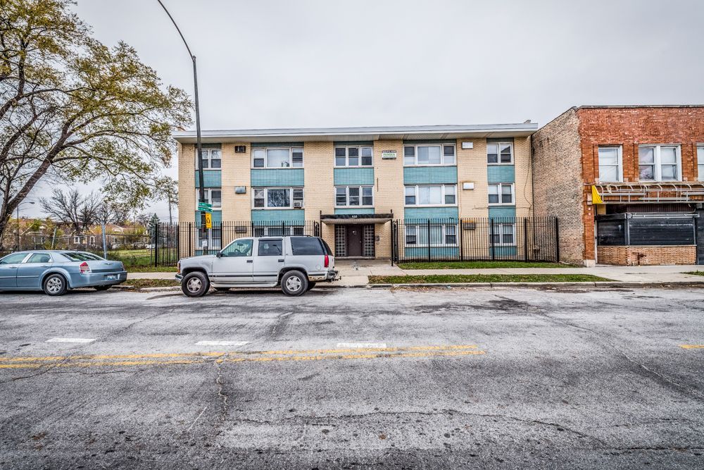 Apartment building with blue trim, parked cars, and adjacent brick building on a city street.