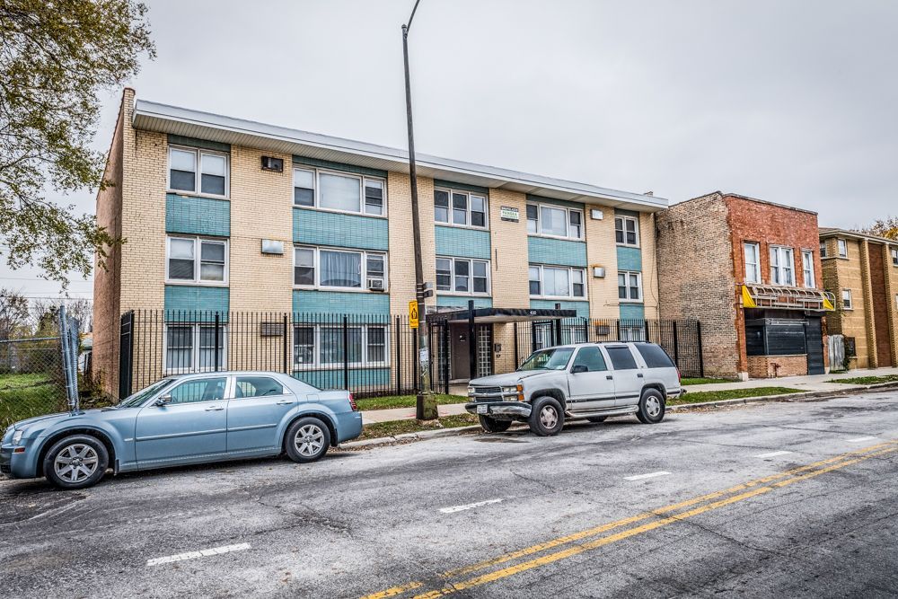 Apartment building on a city street, two cars parked out front, overcast sky.