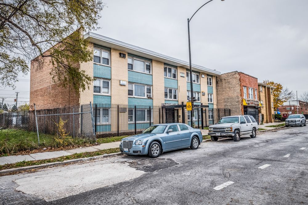 Two-story building with tan, blue, and brick facade. Cars parked on the street. Overcast day.