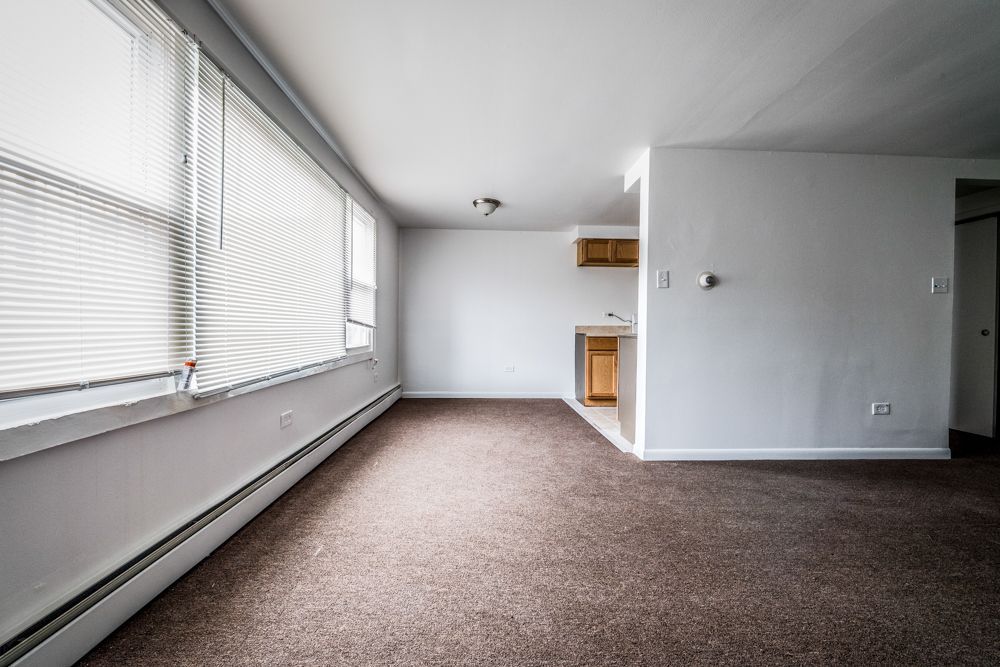 Empty apartment living room with blinds, brown carpet, and small kitchen entrance.