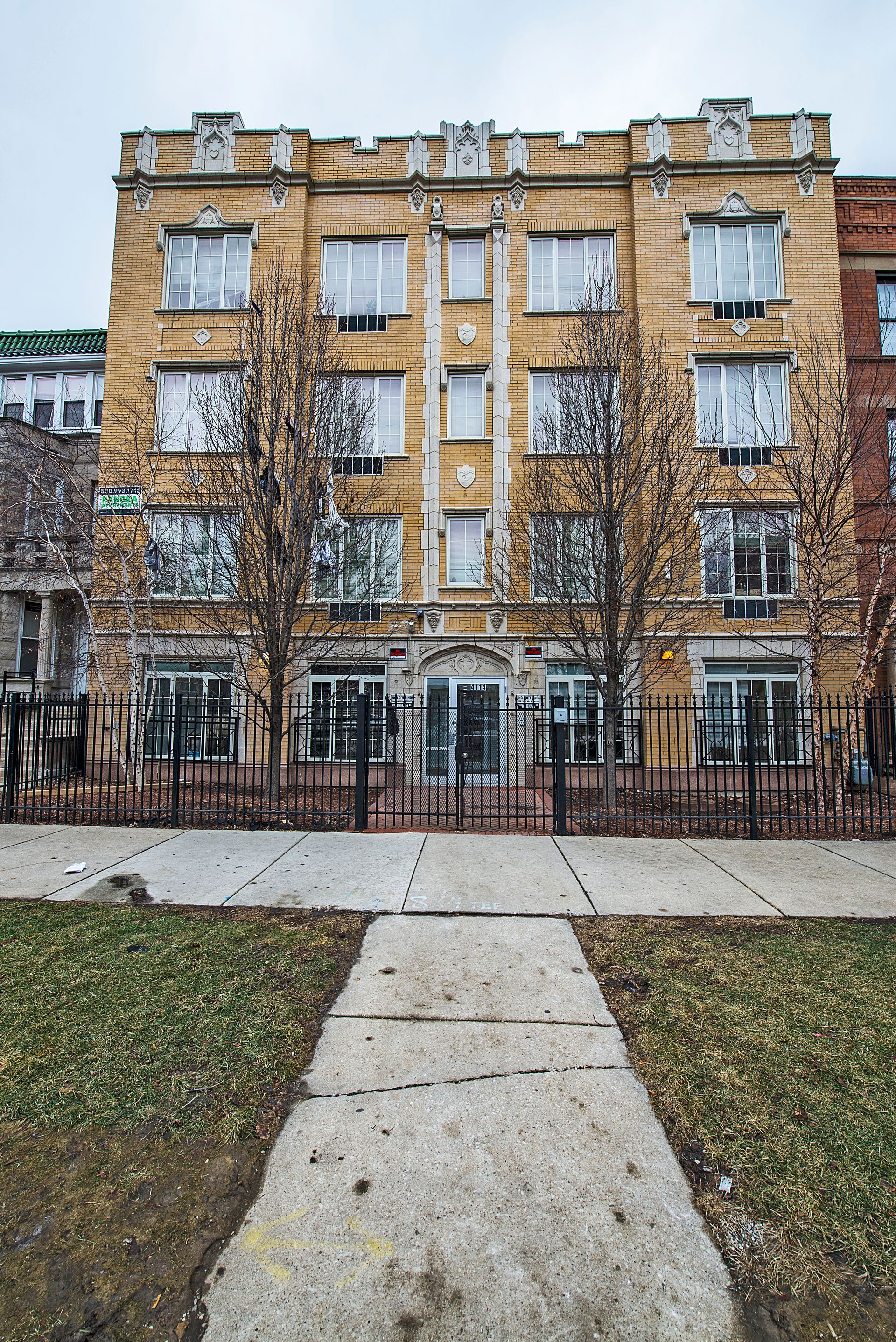 Yellow brick apartment building with black wrought iron fence and sidewalk.