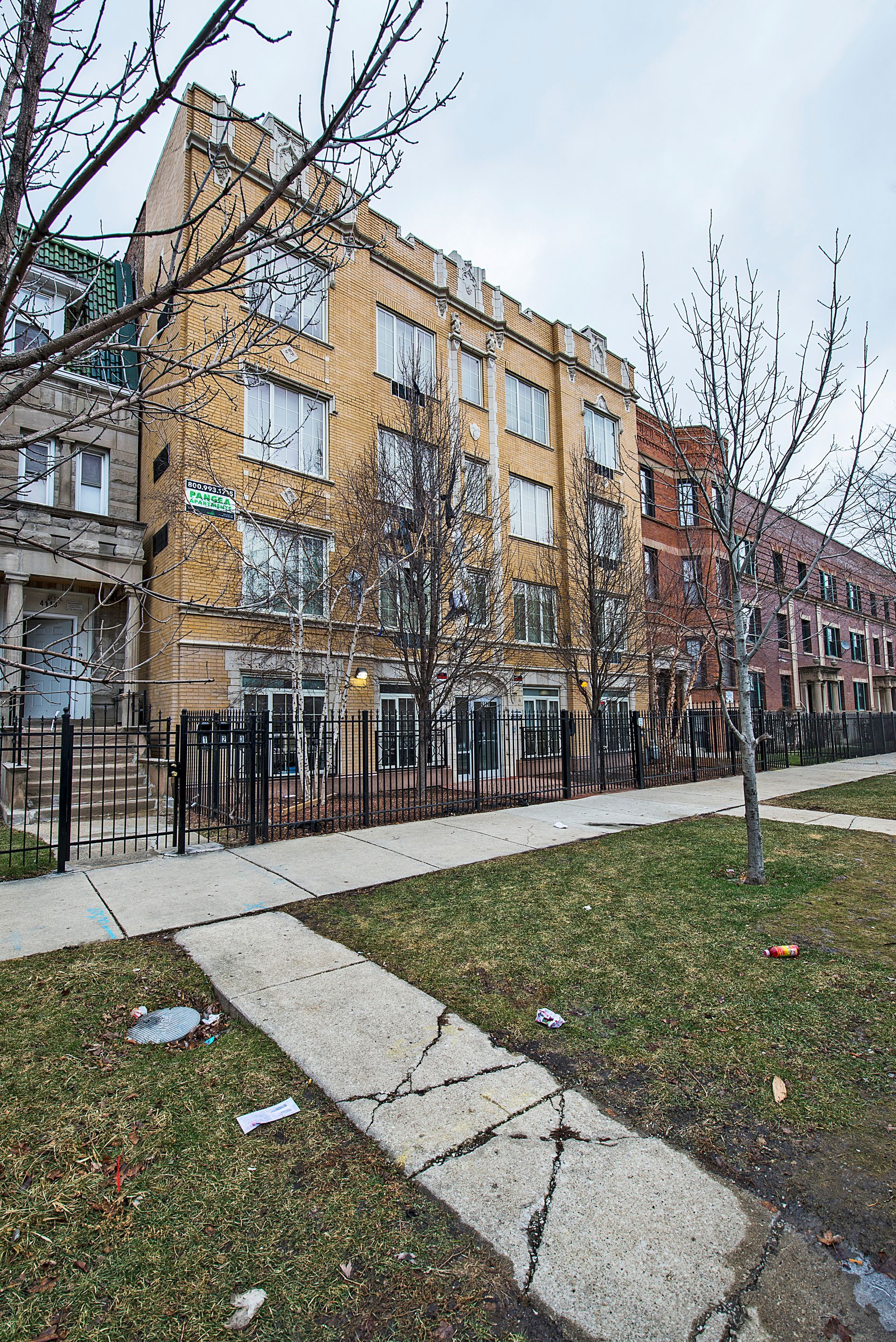 Yellow brick apartment building with black fence, trees, and cracked sidewalk.