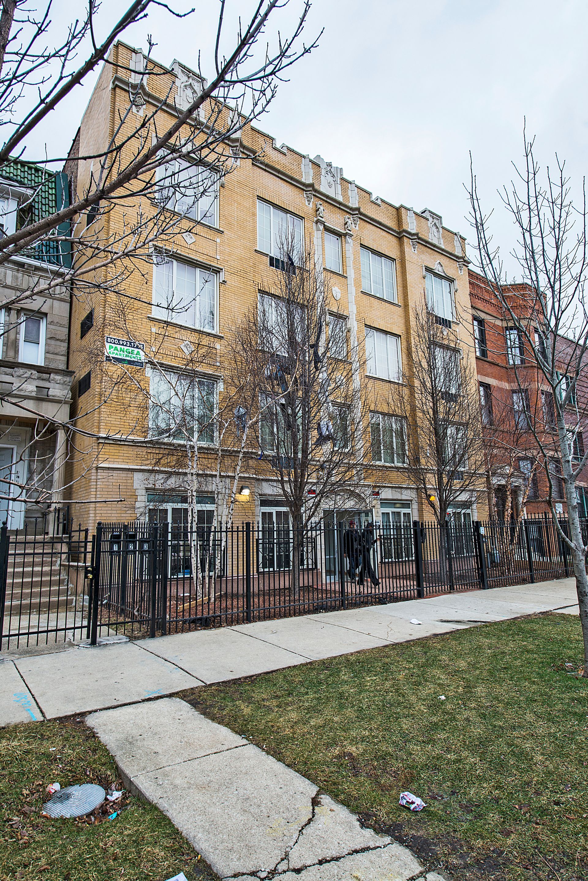 Multi-story yellow brick building with windows behind a black fence. Bare trees in front.