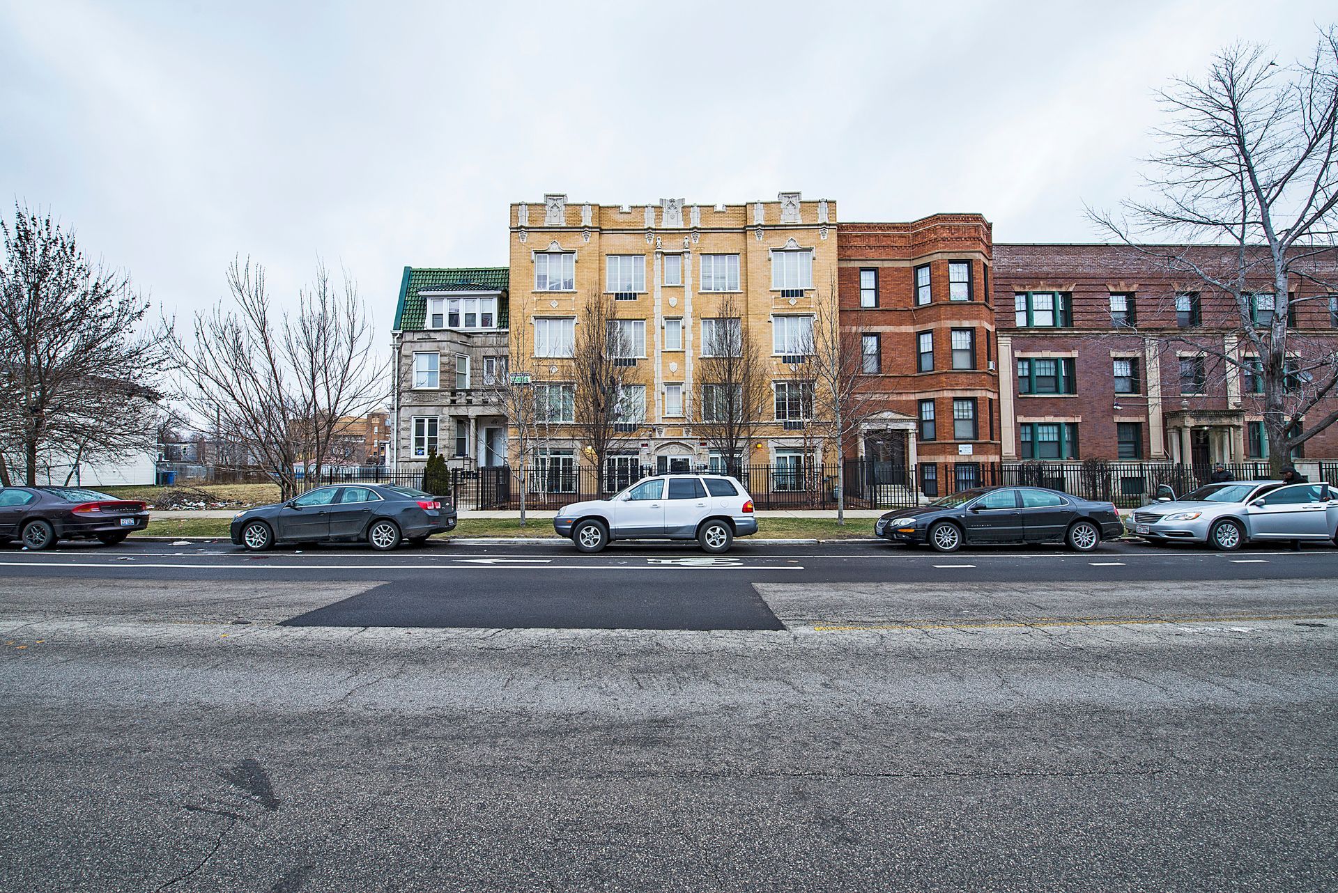 Row of colorful brick buildings with parked cars on a gray street under a cloudy sky.