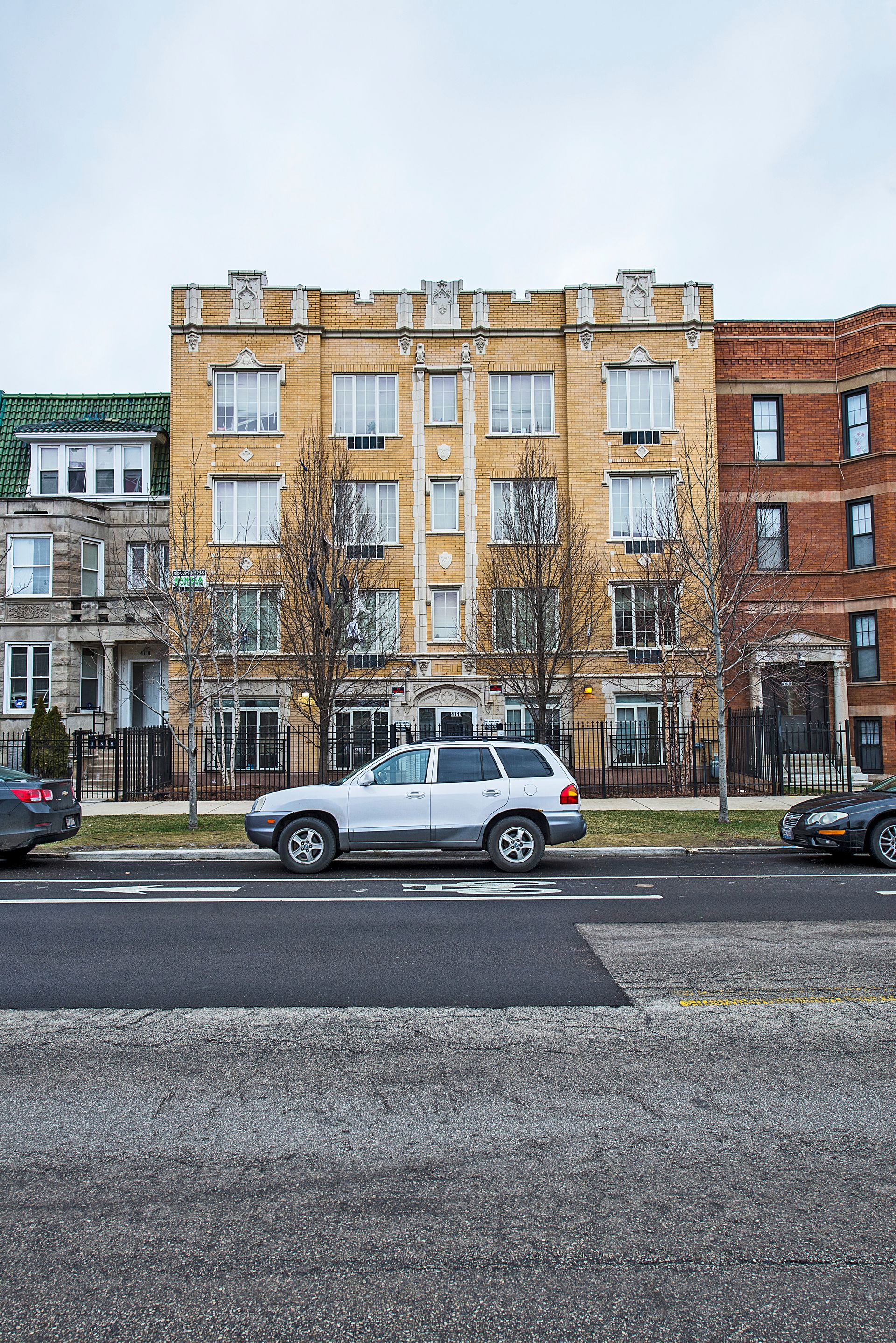 Yellow and brown apartment buildings line a street with a silver SUV parked in front.