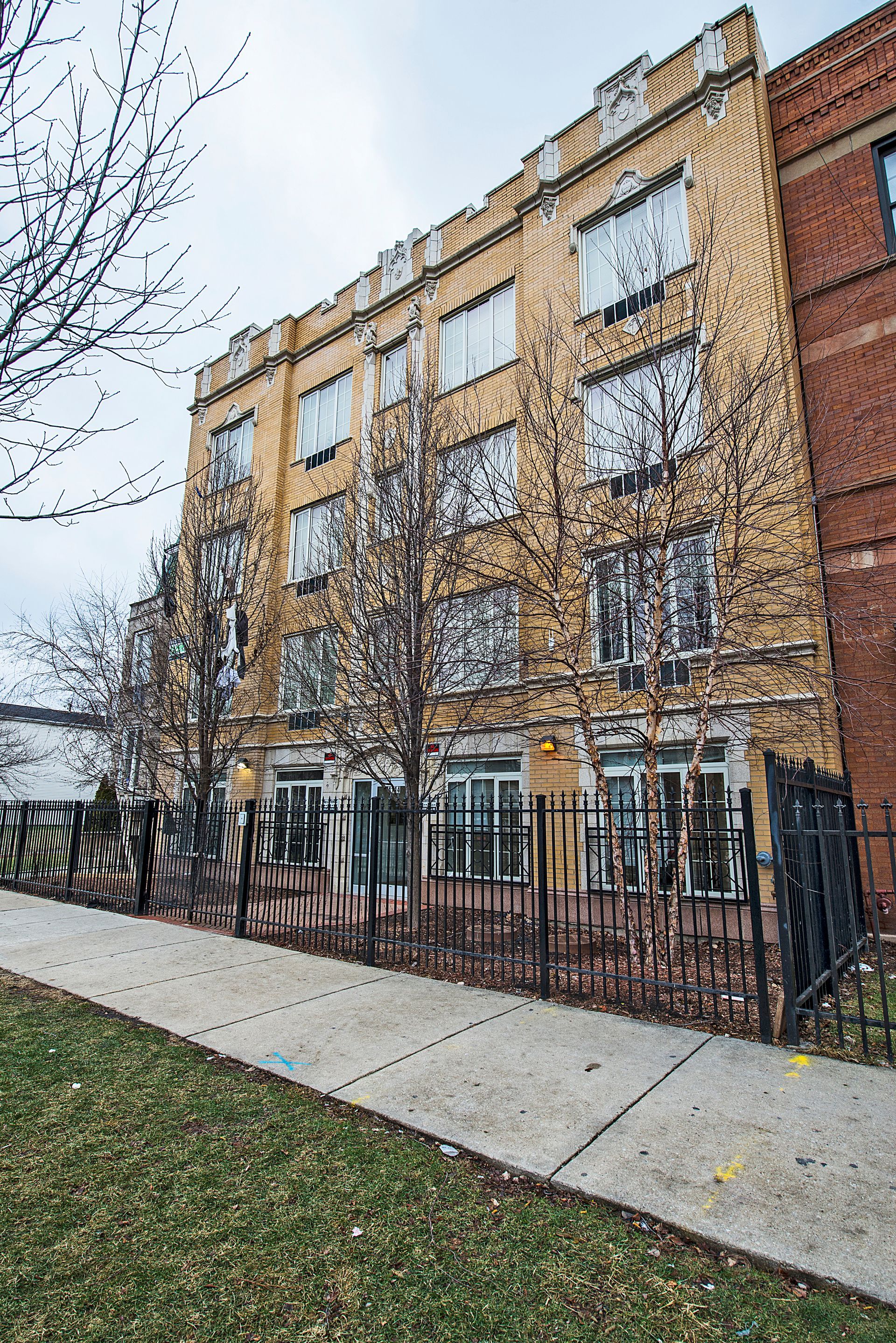 Yellow brick apartment building with black wrought-iron fence and sidewalk. Bare trees and overcast sky.