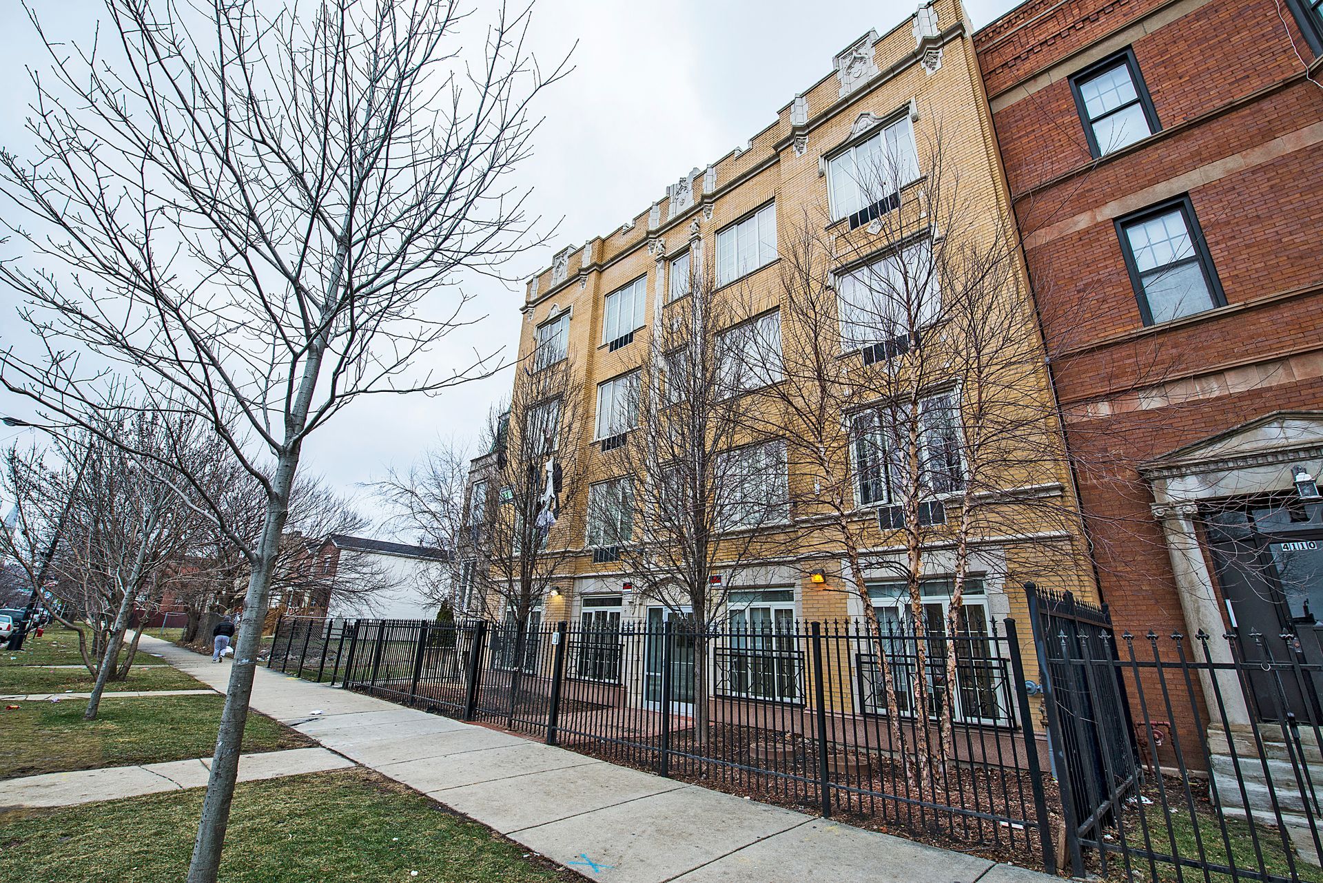 Apartment building with yellow facade, brick building to the right, sidewalk, bare trees.