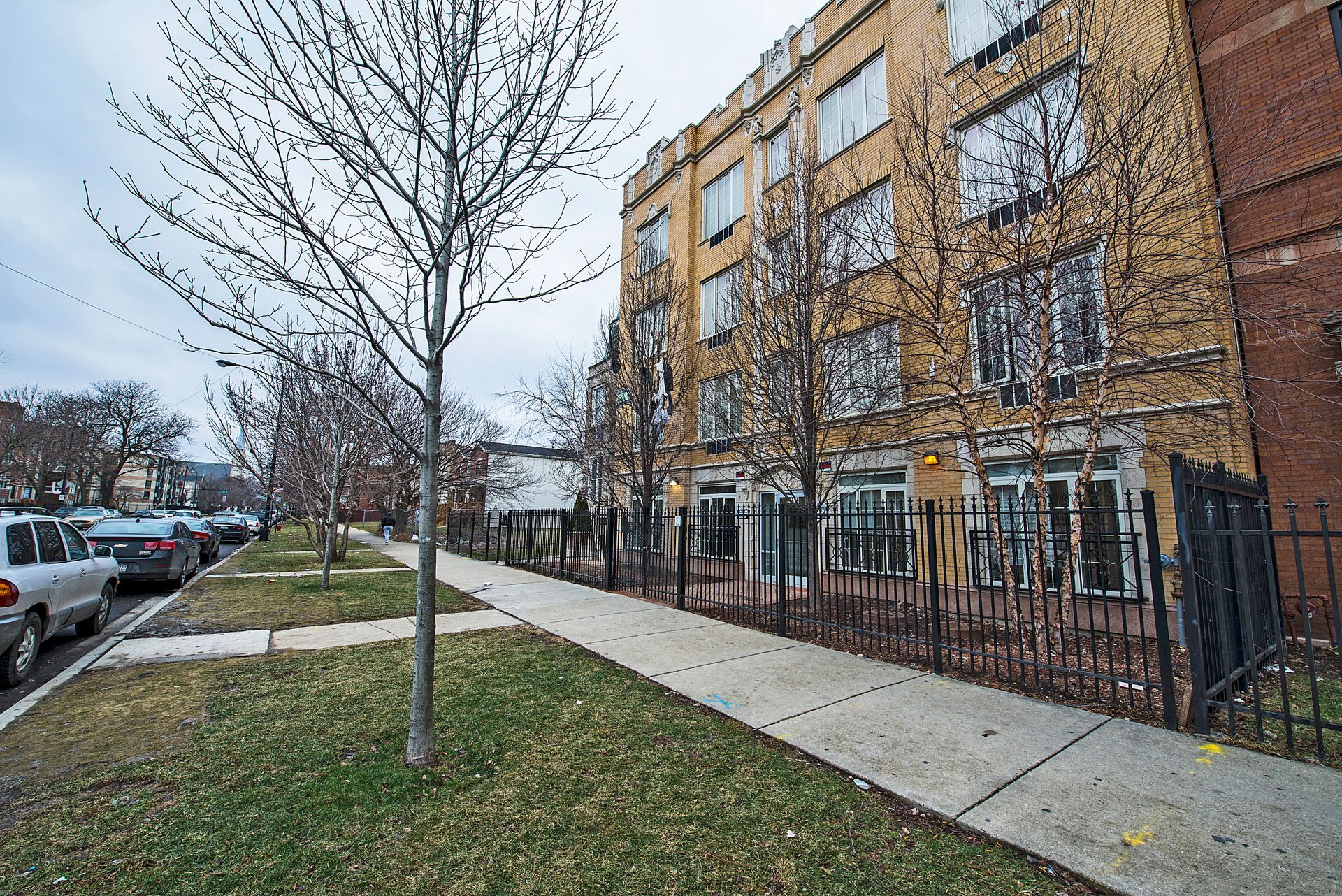 Apartment building on a city street, sidewalk, parked cars, leafless trees, overcast sky.