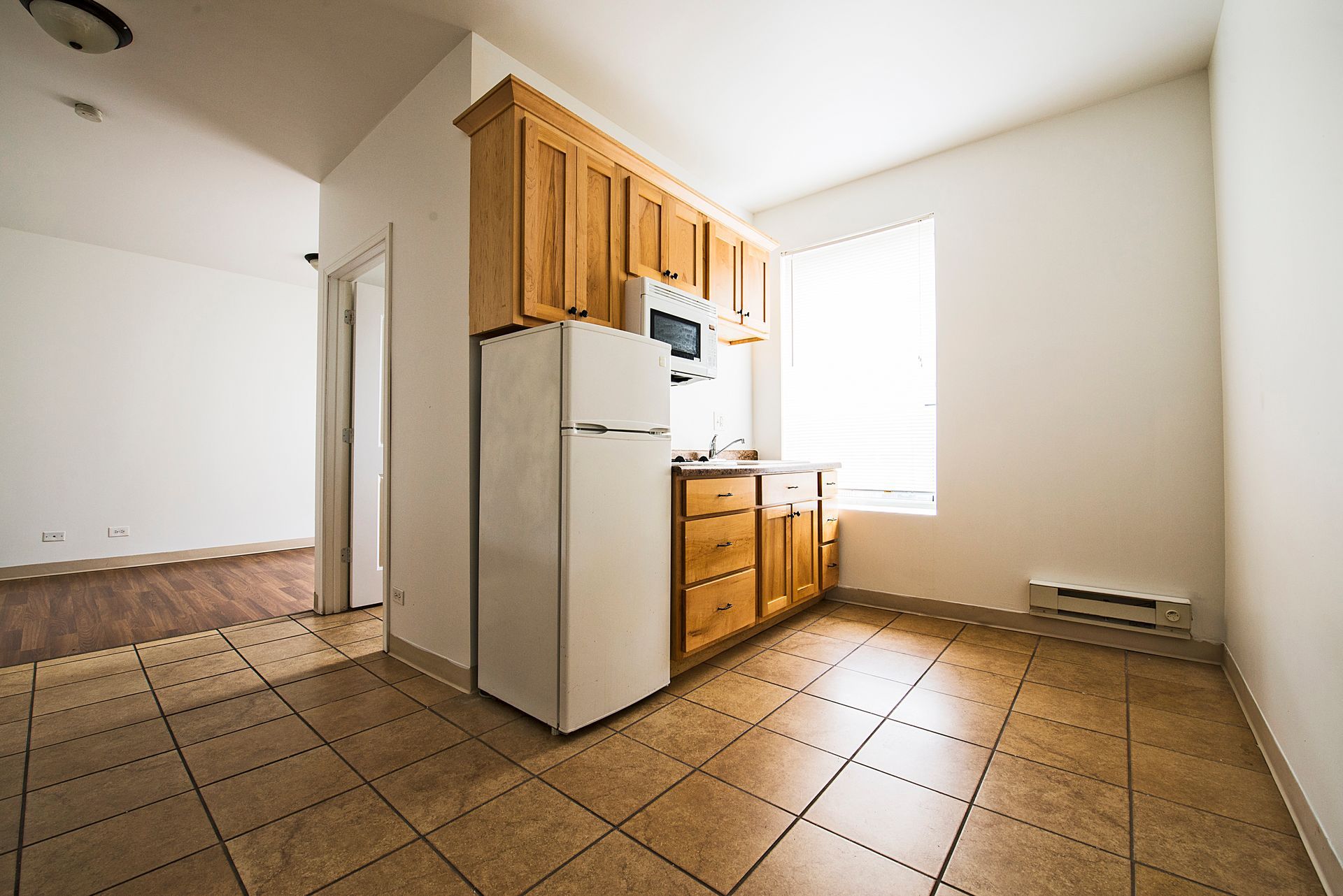 Small kitchen with wood cabinets, refrigerator, and tile floor; doorway to empty room.