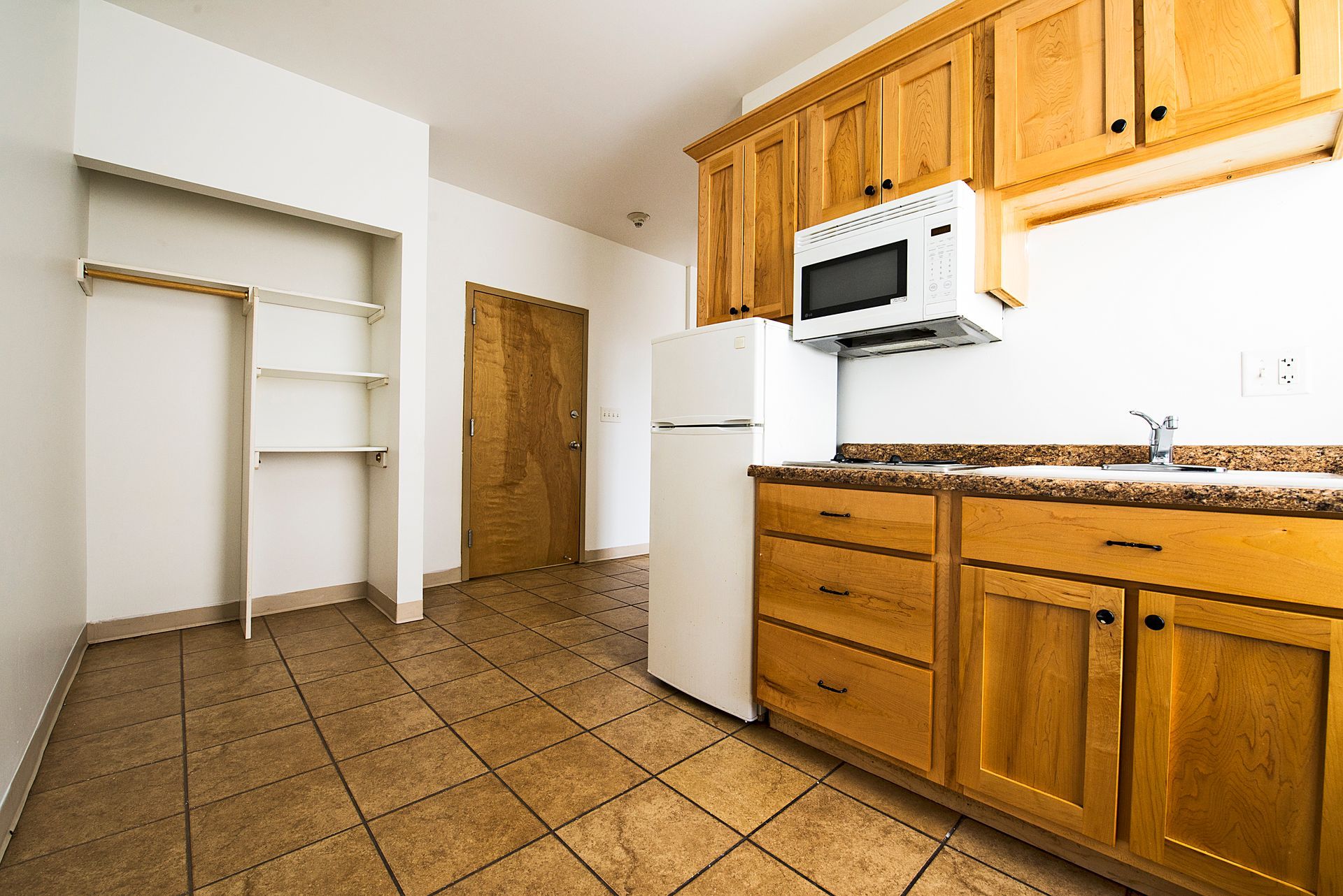 Small kitchen with light wood cabinets, microwave, refrigerator, and tile floor. Closet on the left.