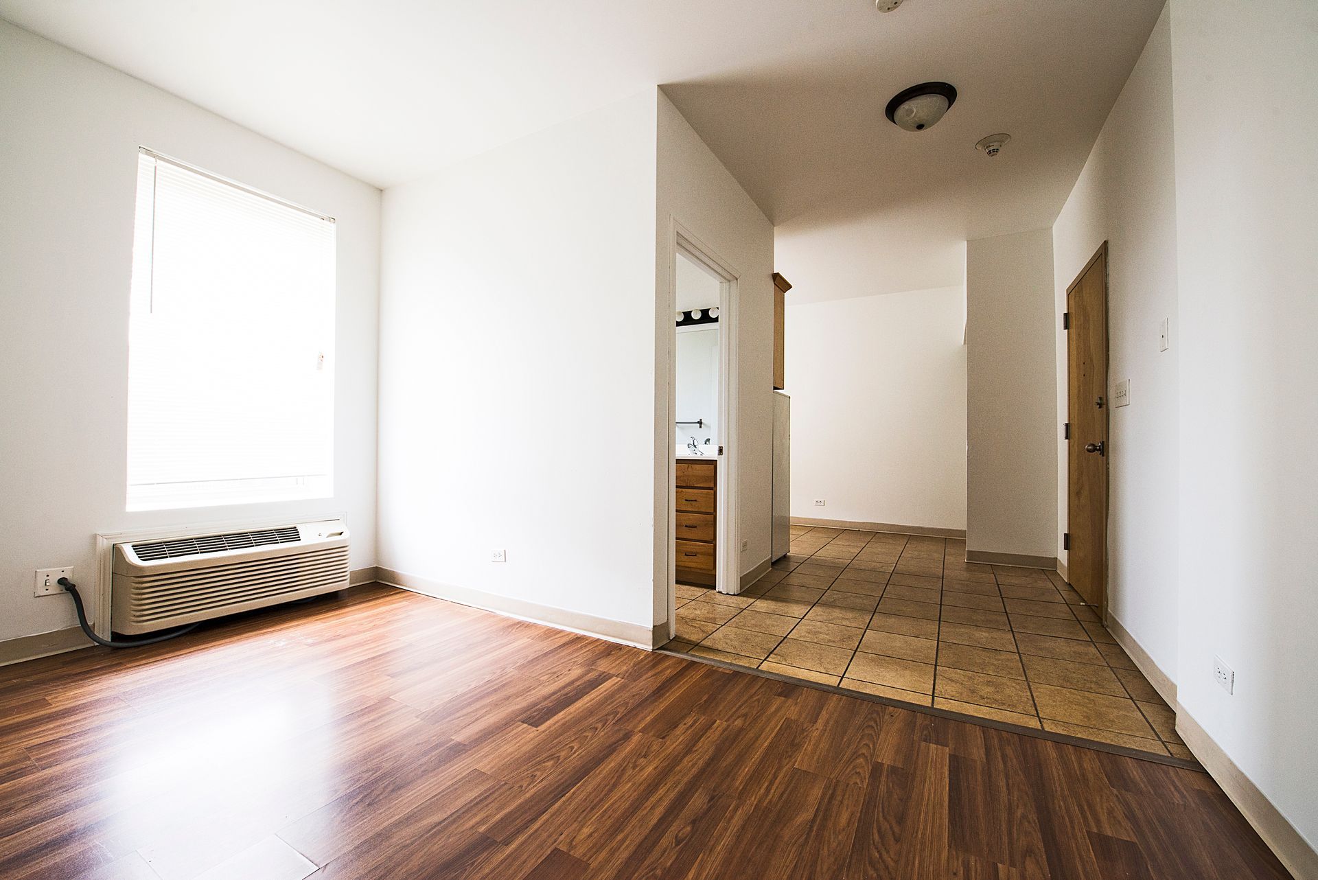 Empty apartment interior with wood flooring, white walls, and a hallway leading to other rooms.