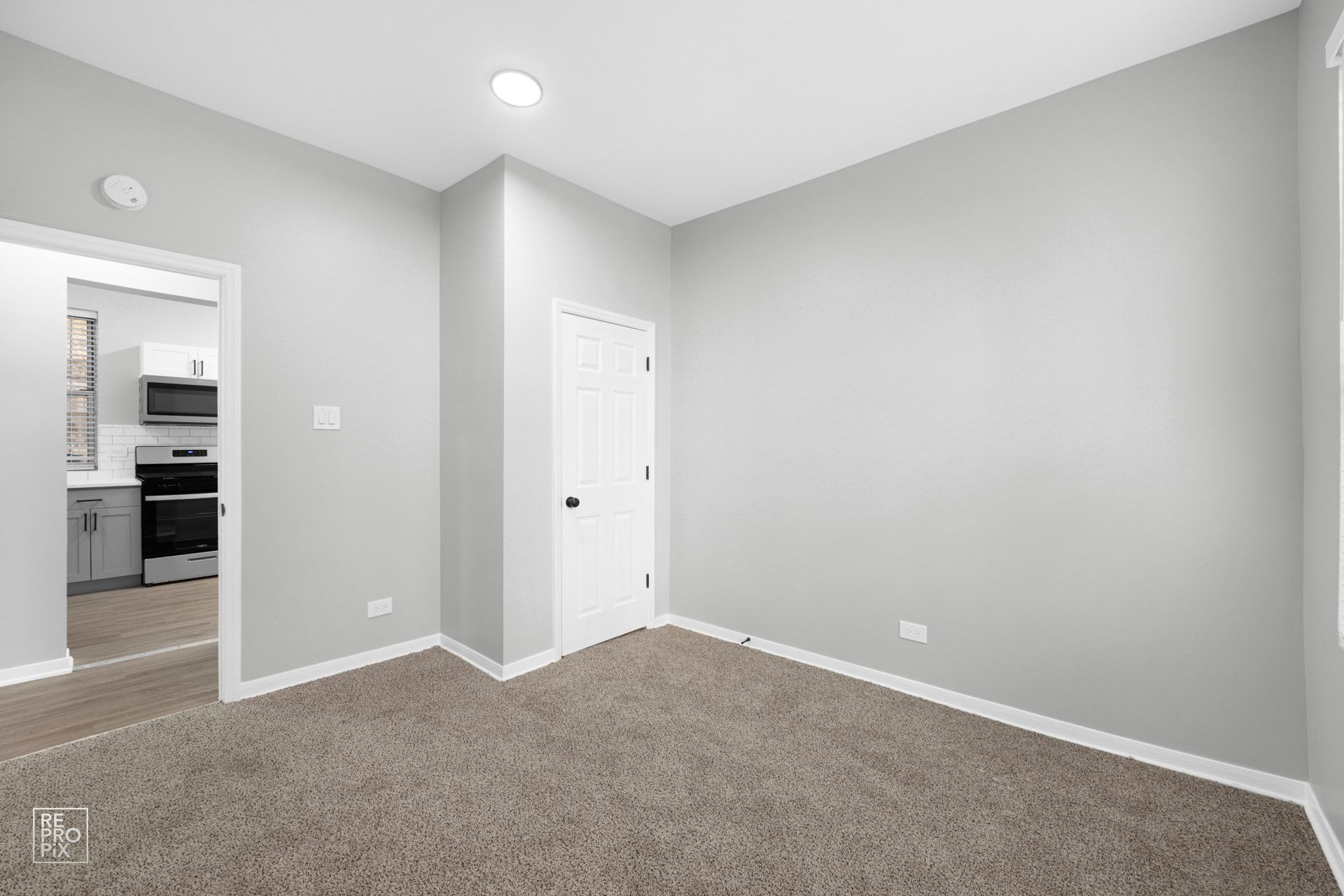 Empty room with grey walls, beige carpet, white door, and doorway to a kitchen.