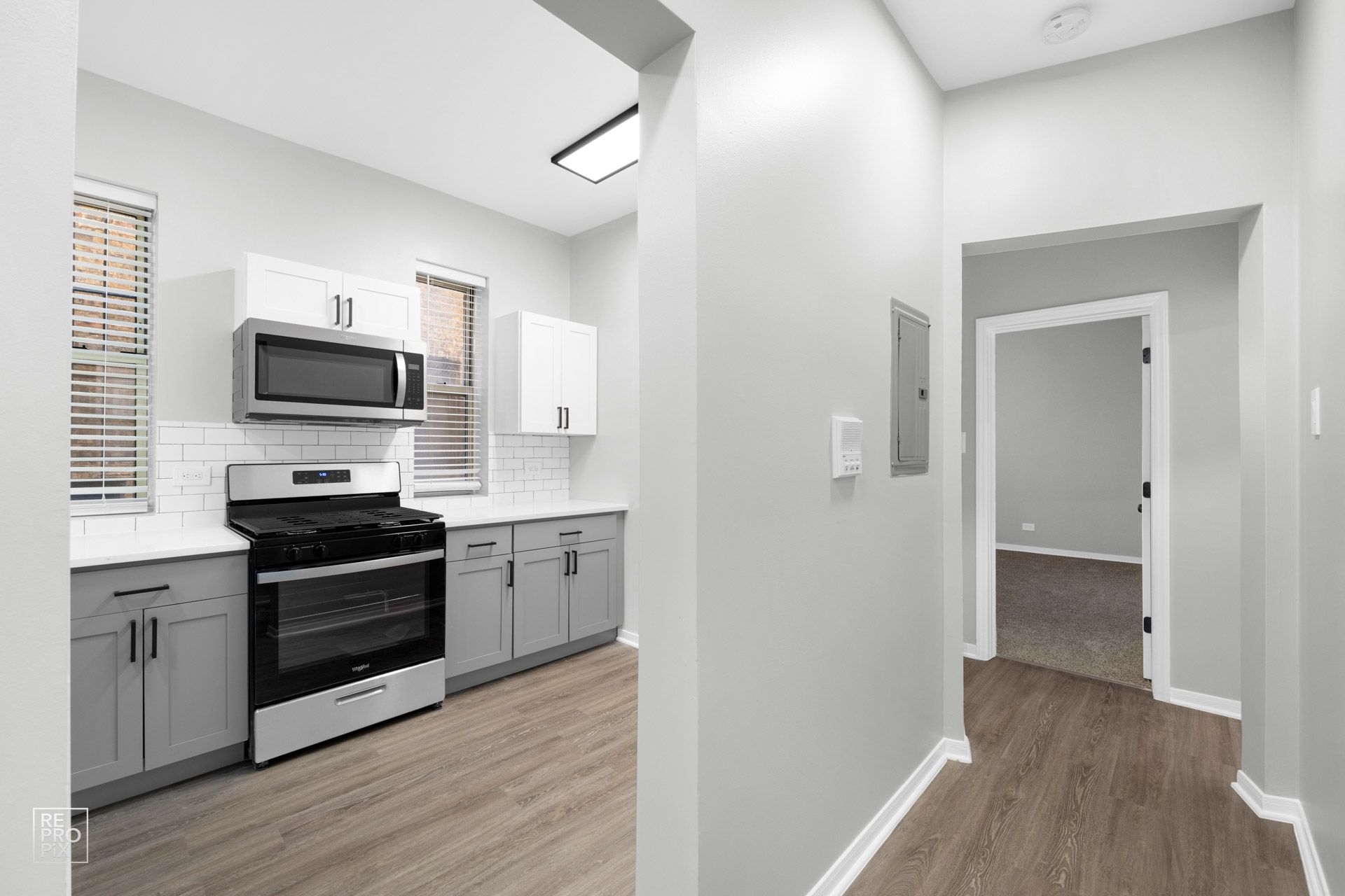 Kitchen with gray and white cabinets, black oven, and light wood flooring. A hallway leads to a room.