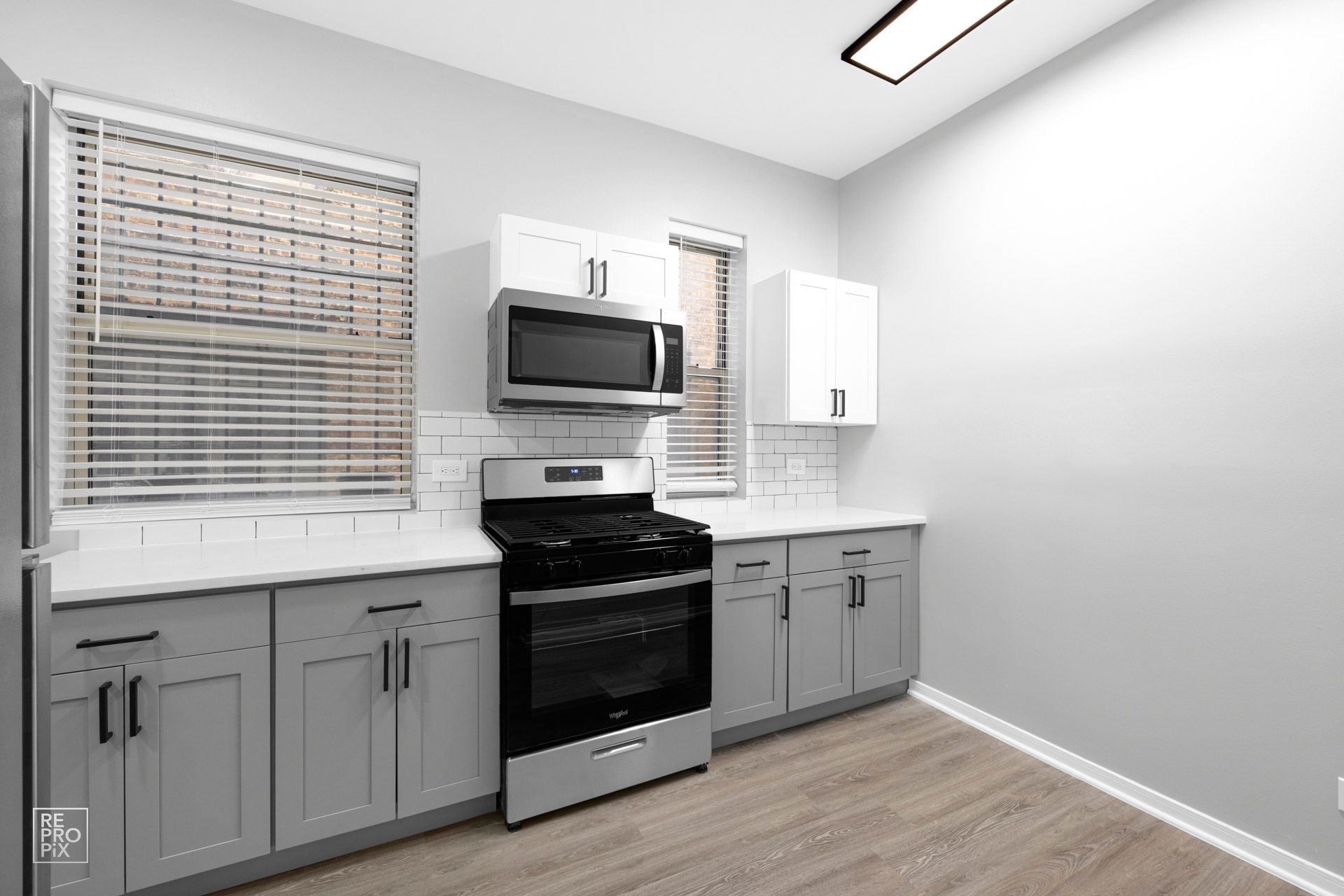 Gray and white kitchen with stove, cabinets, microwave, and windows with blinds.