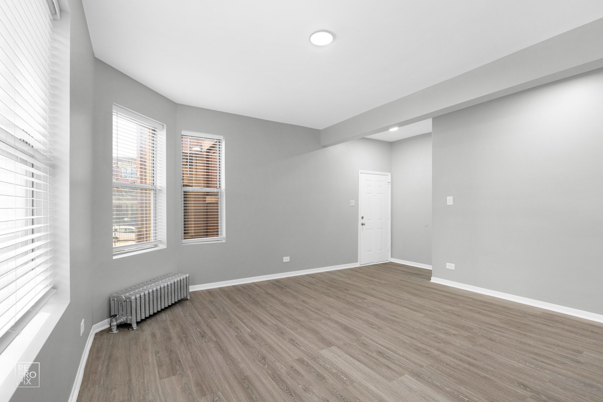 Empty room with gray walls, wood-look flooring, windows with blinds, and a door.