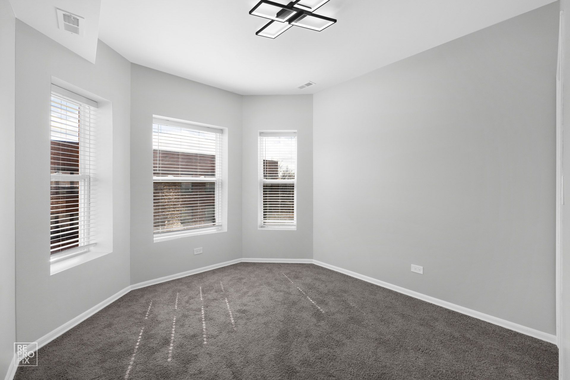 Empty room with gray walls and carpet, three windows, and a modern ceiling light.