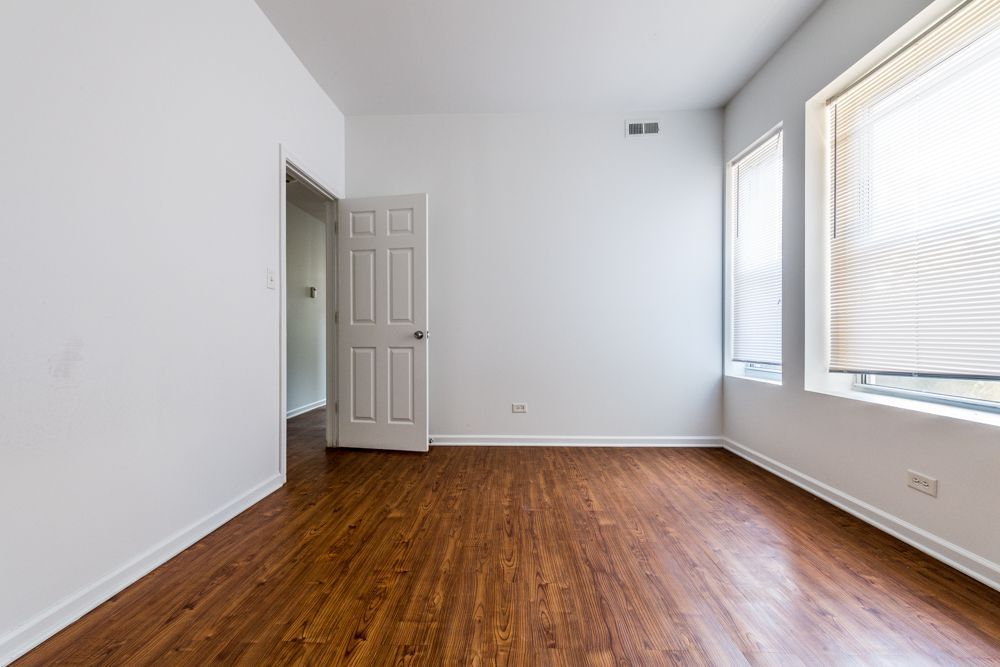 Empty room with hardwood floors, white walls, and a closed door. Windows with blinds.