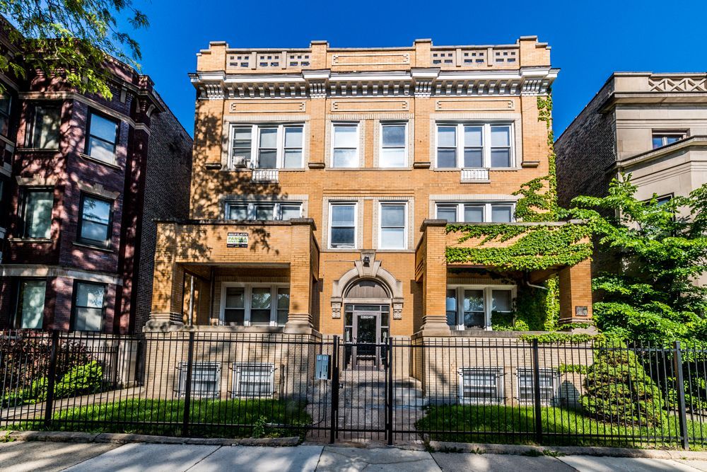 Three-story brick building with arched entry, windows, and wrought iron fence. Green foliage on right.