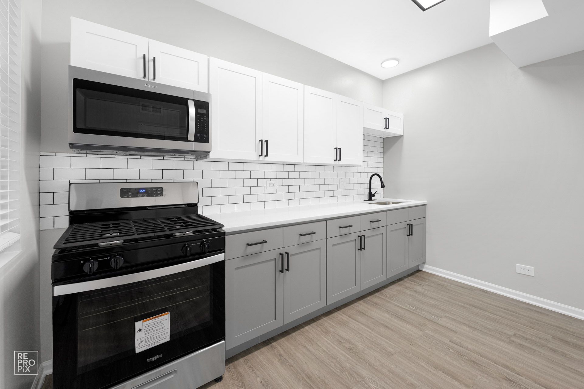Modern kitchen with gray and white cabinets, stainless steel appliances, and white tile backsplash.