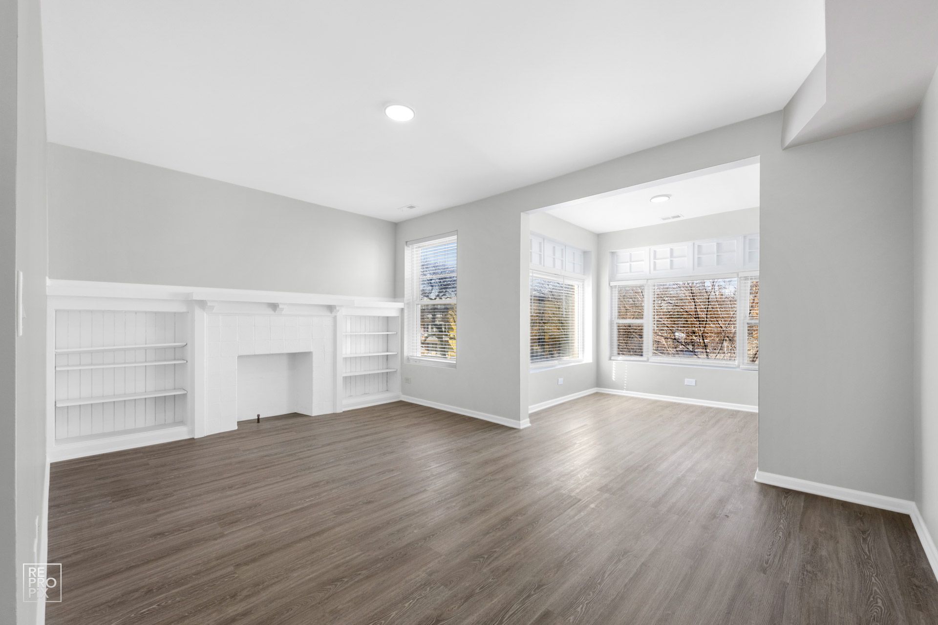 Empty living room with gray walls, built-in shelving, dark wood-look floor, and three windows.