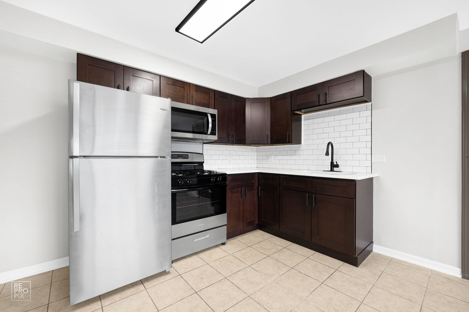 Kitchen with stainless steel appliances, dark cabinets, white backsplash, and tan tile floor.