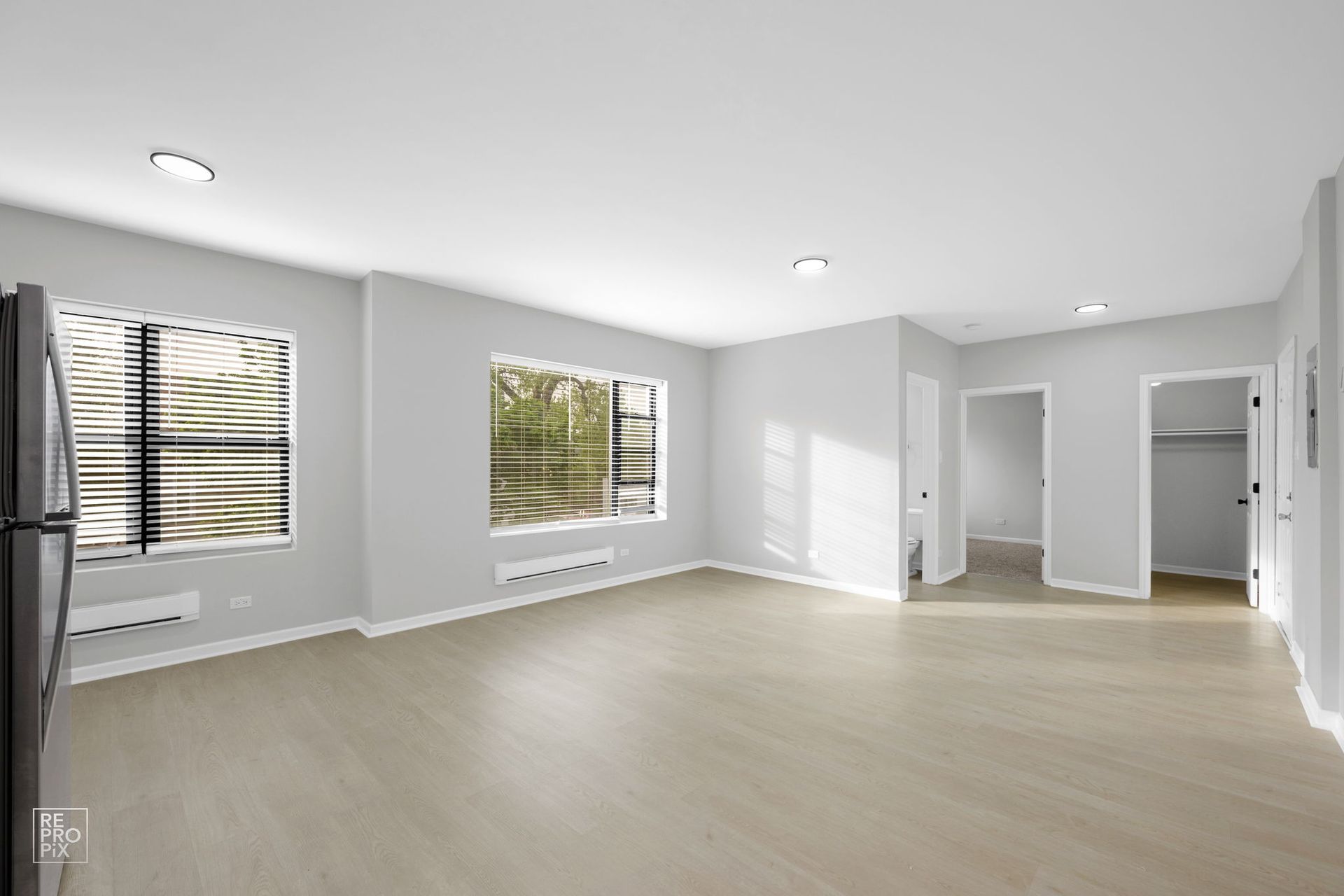 Empty, bright living room with large windows, light wood floors, and neutral gray walls.