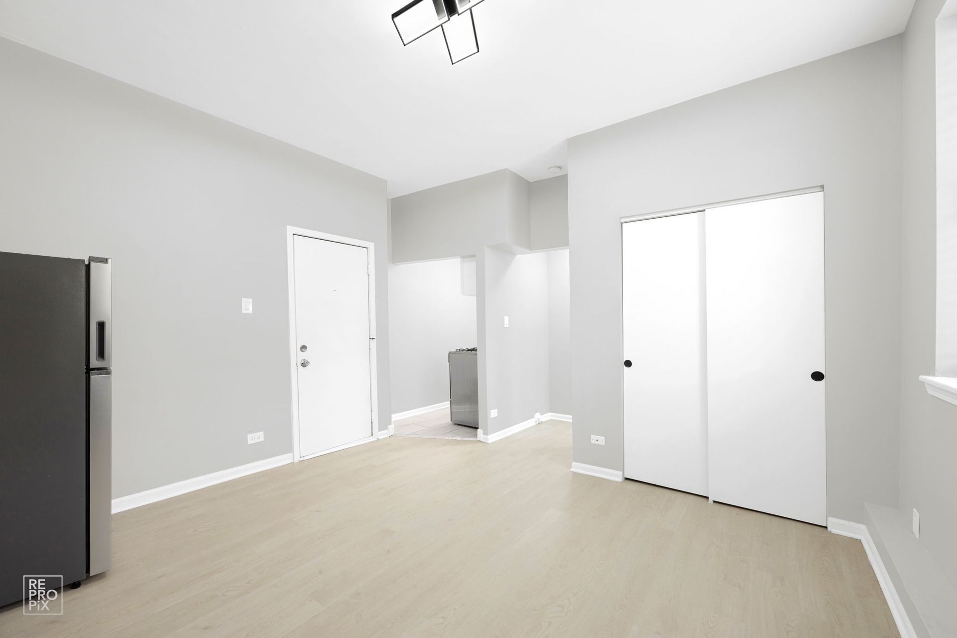 Empty, light gray-walled apartment with white doors, refrigerator, and ceiling light.