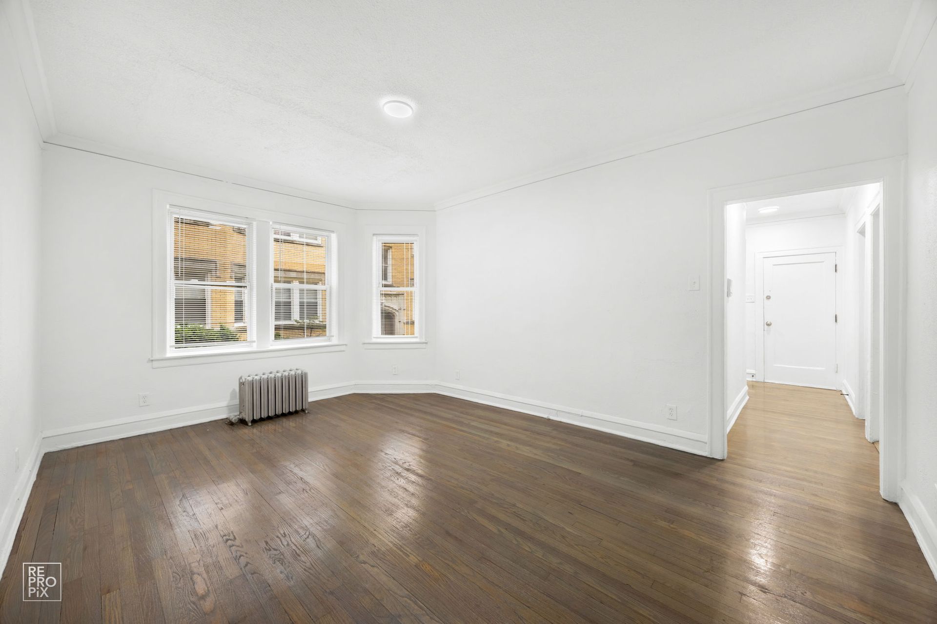 Empty, sunlit room with hardwood floors, white walls, and a hallway doorway.