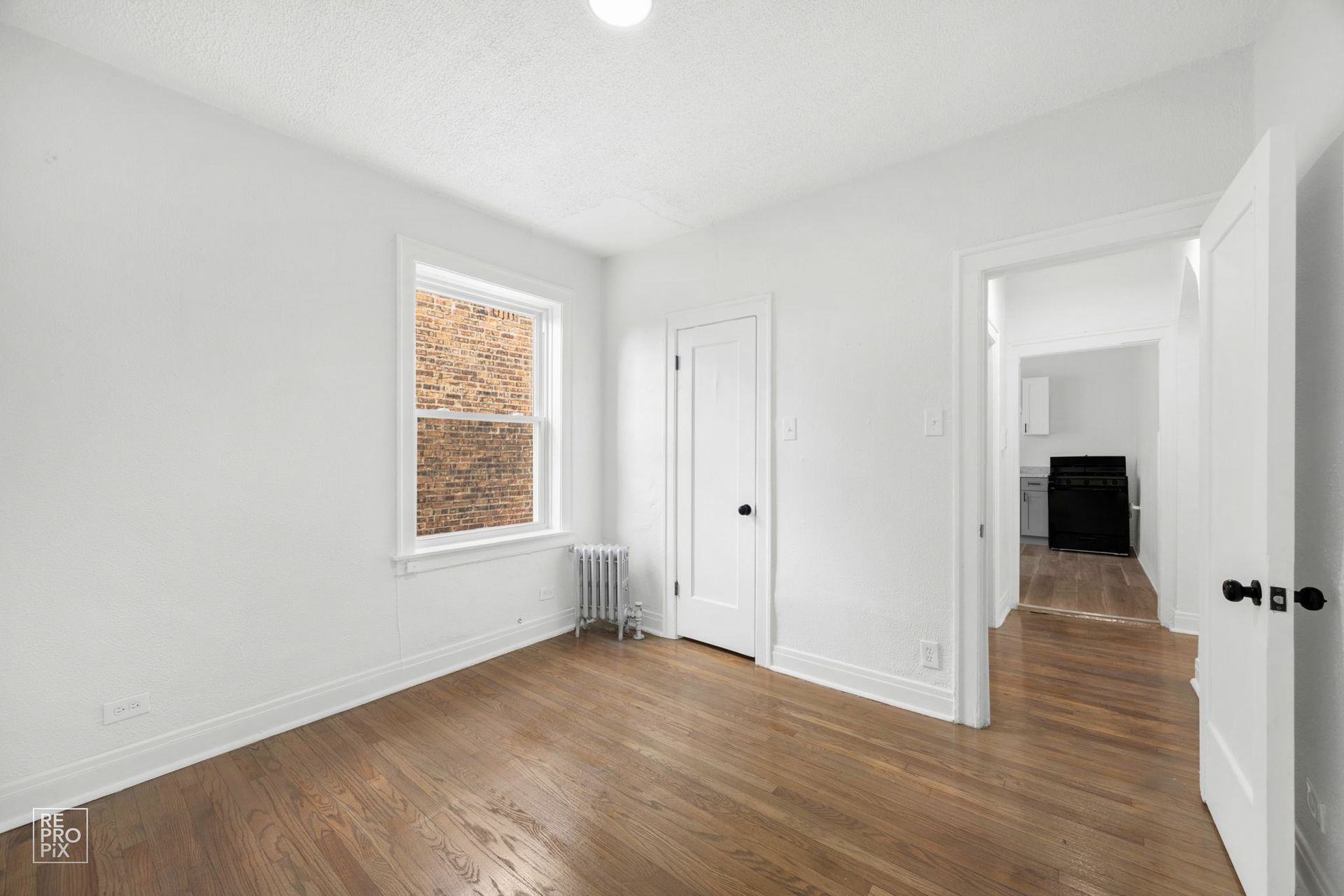 Empty bedroom with hardwood floors, white walls, window, door, and doorway to another room.