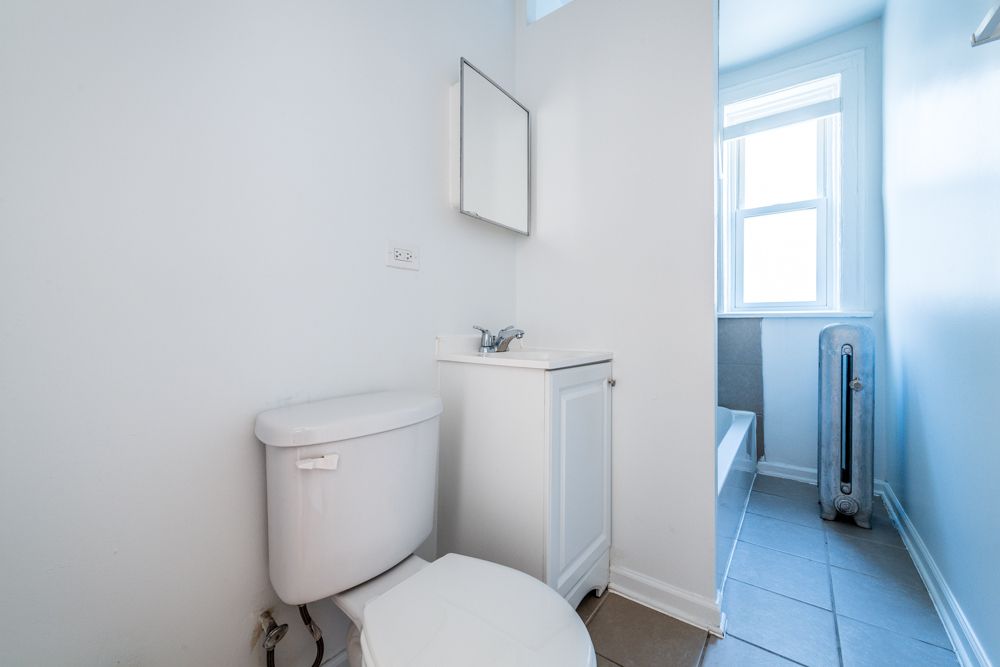 Small white bathroom with toilet, sink, and window with radiator.