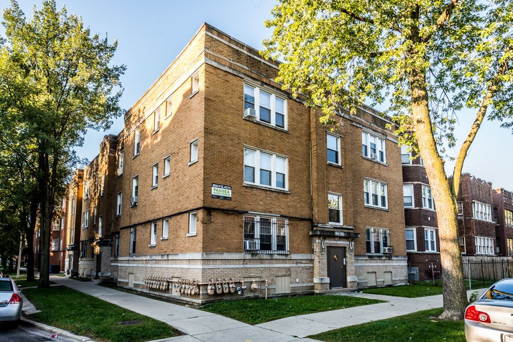 Brick apartment building with multiple windows, trees, and cars parked on the street.