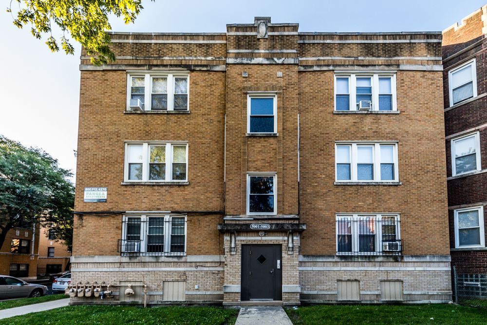 Three-story brick apartment building with white-trimmed windows and a dark door, sidewalk in front, green grass.