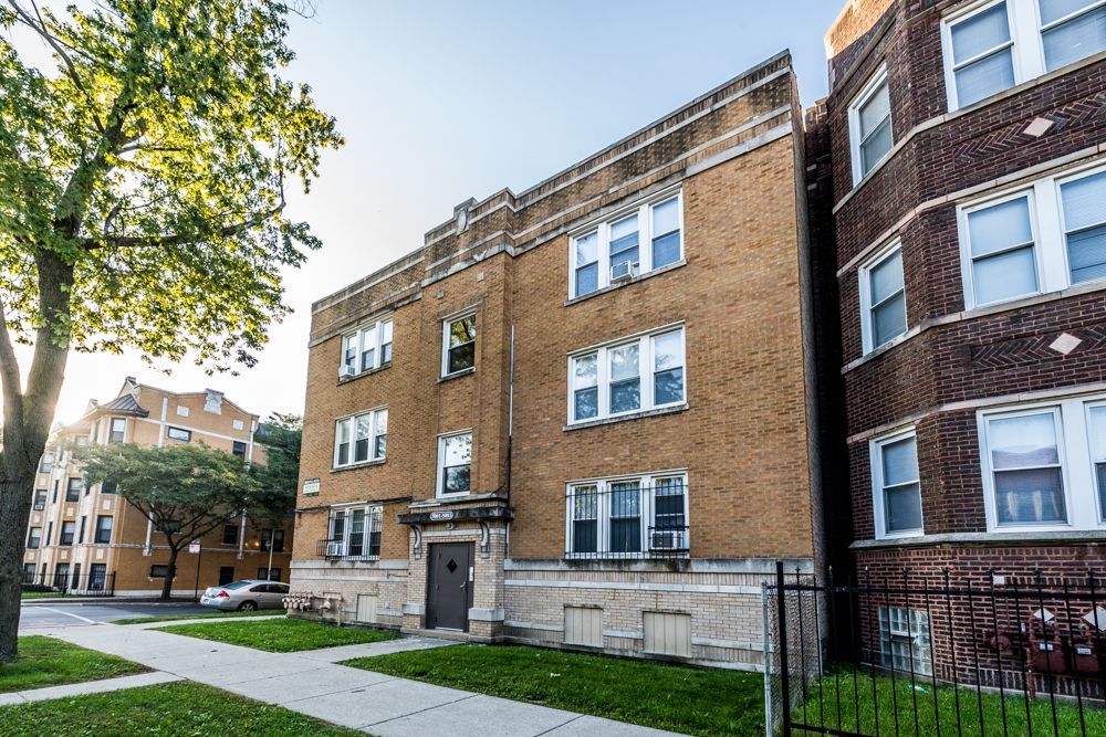 Three-story brick apartment building on a residential street; a sidewalk and green lawn in front.