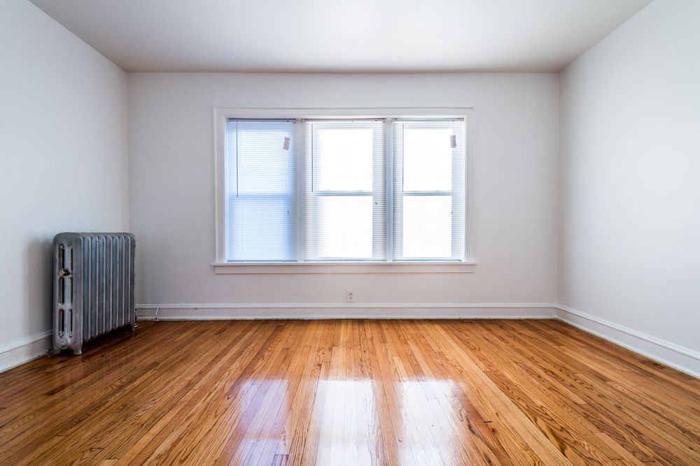 Empty room with hardwood floors, radiator on the left, and three-pane window with blinds.