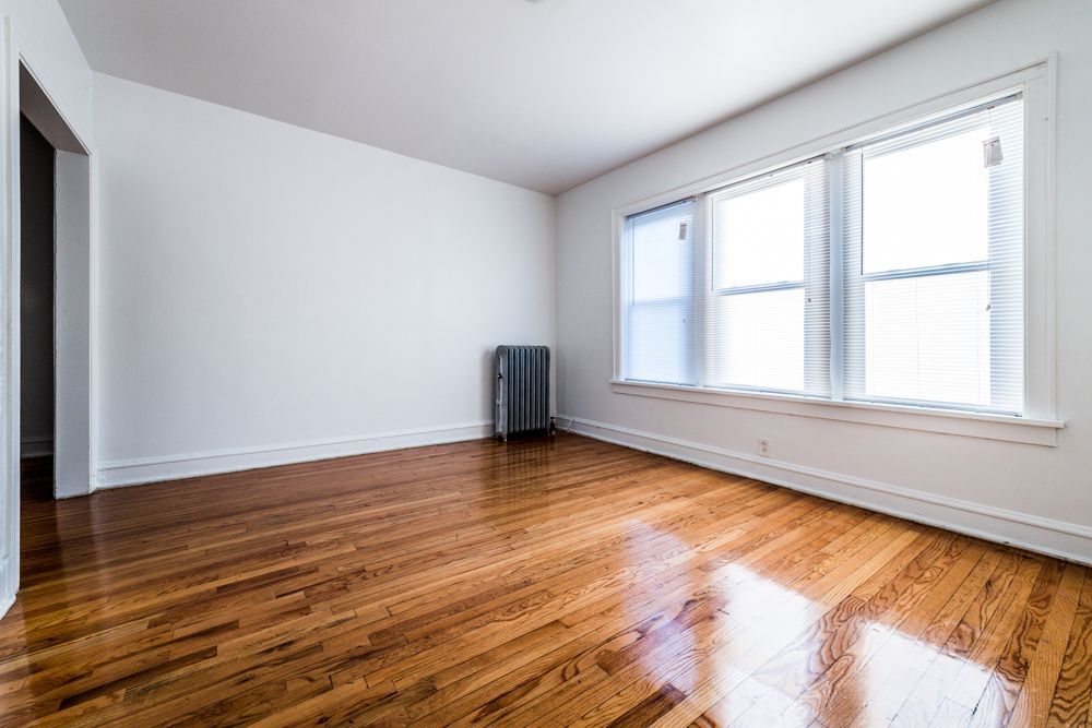 Empty room with hardwood floors, white walls, and a radiator. Three windows provide natural light.