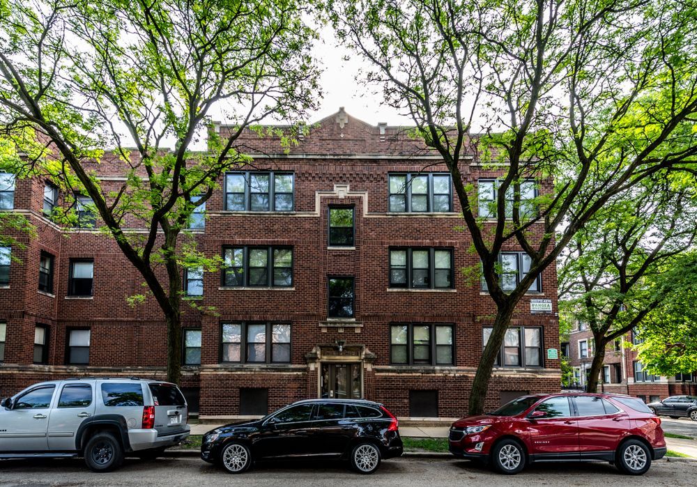 Red brick apartment building with parked cars and trees lining the street.