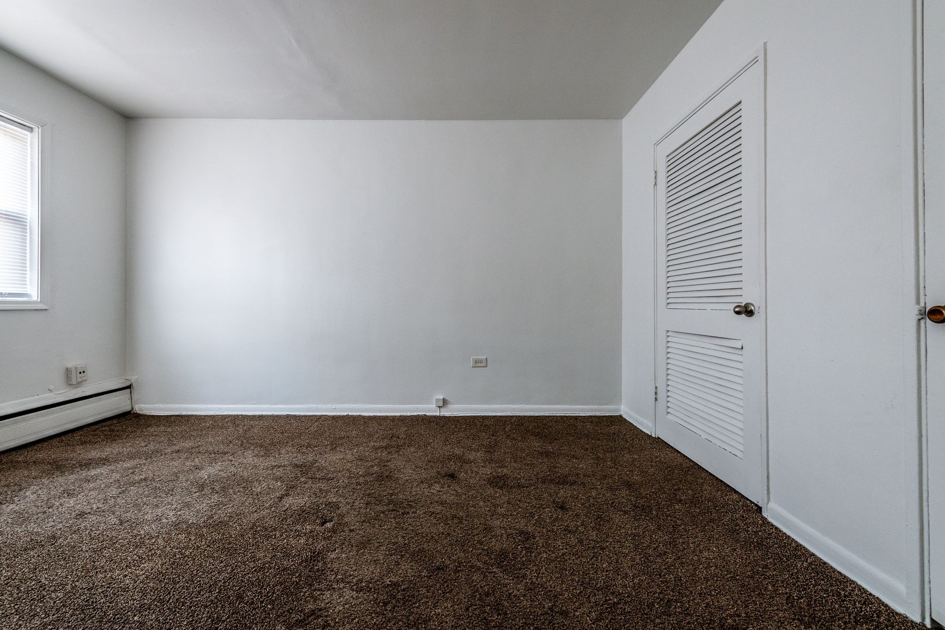 Empty room with white walls, brown carpet, and a white closet door. A small window is on the left.