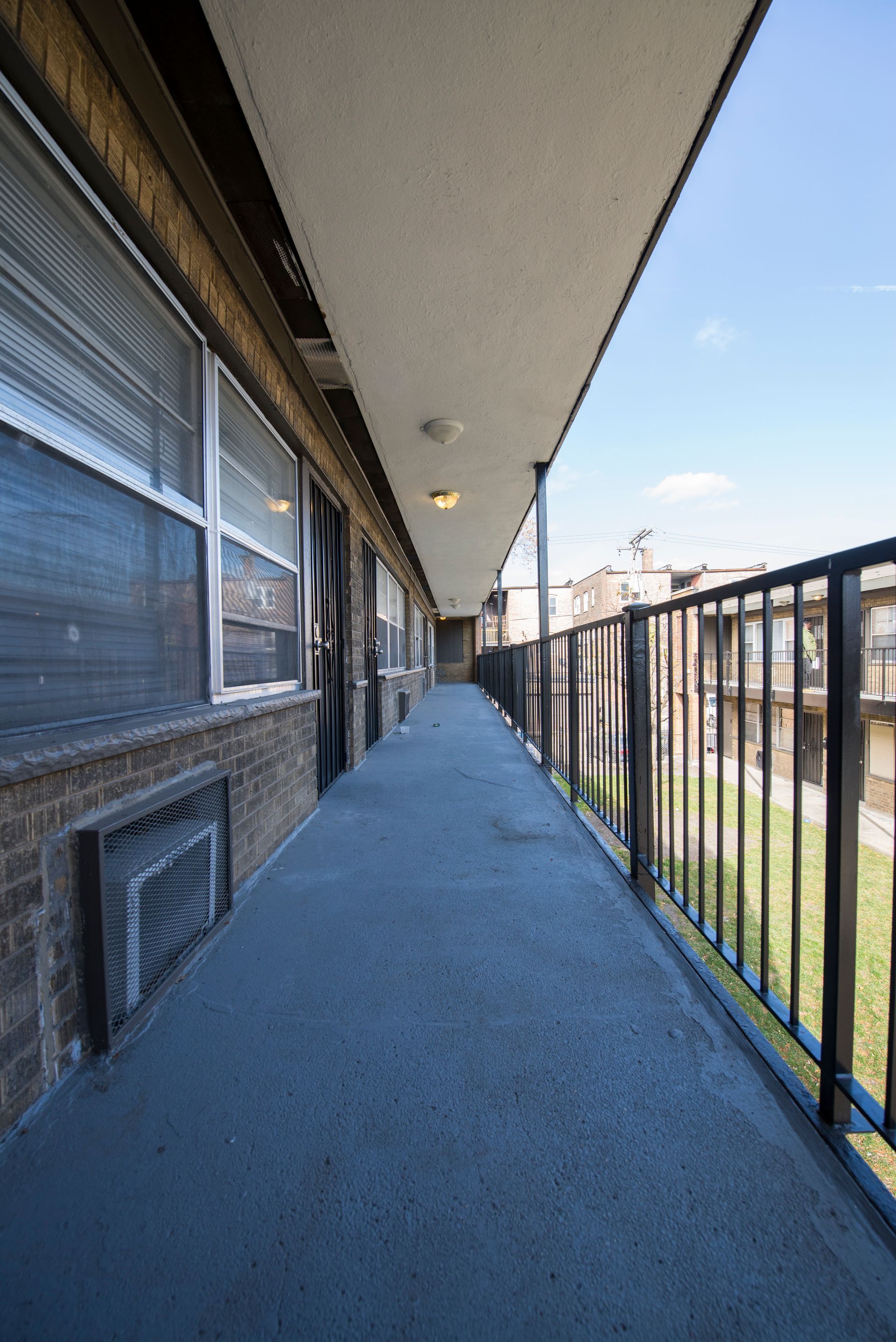 Long exterior hallway with brick wall, windows, and black railing overlooking grass and sky.