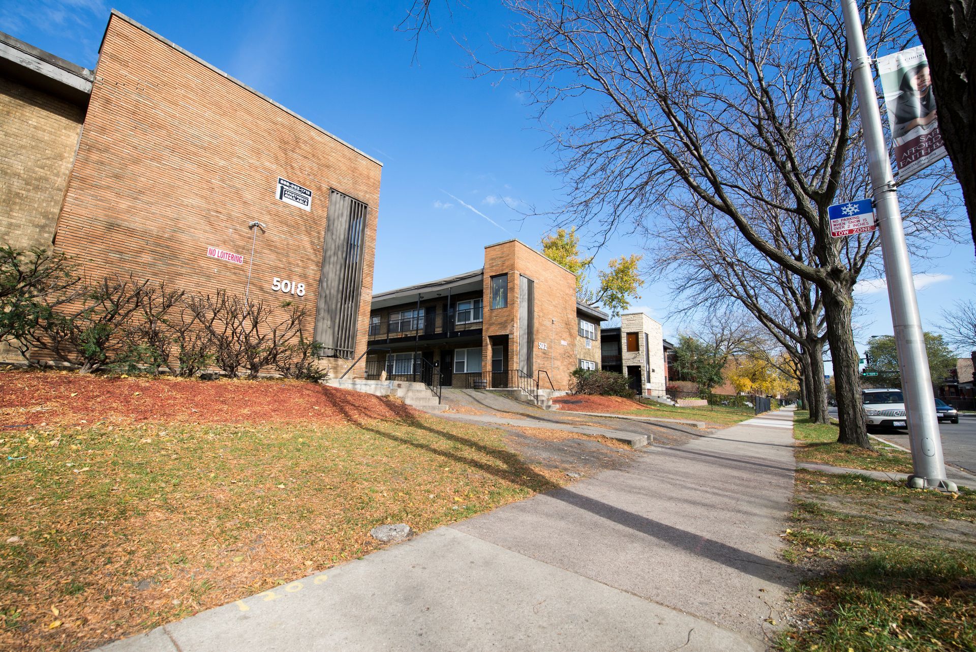 Buildings with a sidewalk in front. Brown brick, autumn leaves. Blue sky.